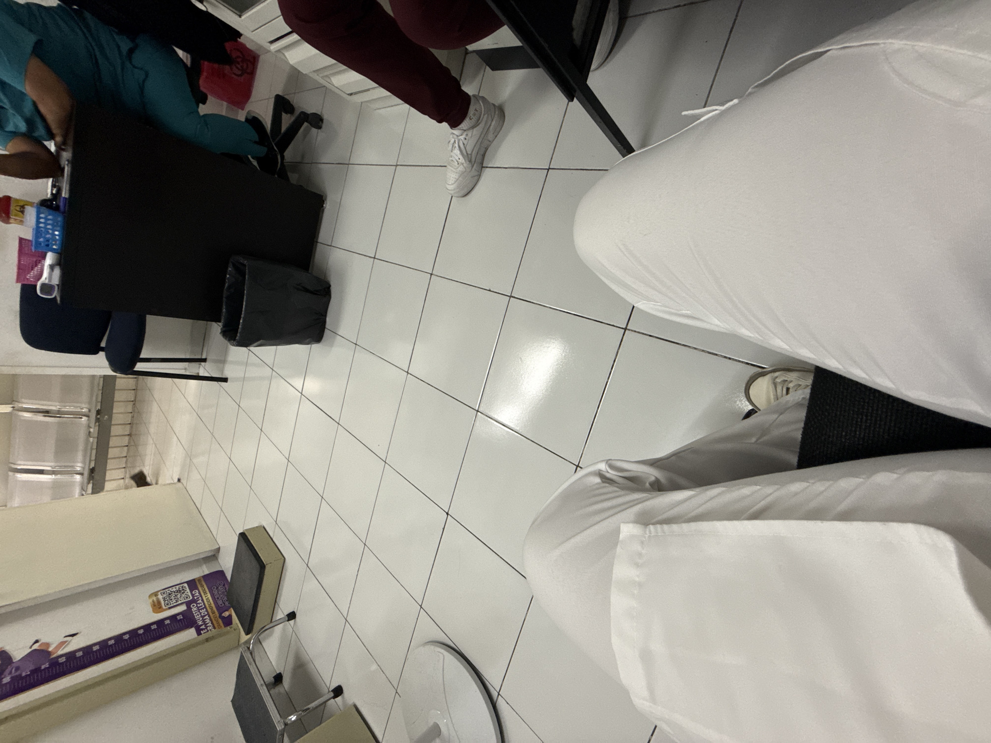 The image captures an indoor scene from a low, angled perspective, likely within a medical or clinical setting. In the immediate foreground on the right, the legs of the photographer, clad in white pants or a garment, are visible. To the left, a person wearing teal scrubs is partially seen, seated at a black desk that holds a blue basket, a white device (possibly a thermometer), and other small items. A black trash can with a liner sits beside the desk, and a red biohazard bag is visible behind it.
Further into the scene, another individual is seated, their lower legs and feet visible, wearing maroon pants and white sneakers. The entire floor is covered in clean white tiles with dark grout lines. In the background on the left, a light-colored shelving unit is present. A purple stadiometer, a device for measuring height, stands upright, displaying a QR code and visible Spanish text that includes "TE A NUESTRO ROGRAMA DE EDAD" (suggesting "Join our age program") and "CLÍNICO AMIGO" ("Friendly Clinic"). A small gray and black stool is situated near the stadiometer. The environment, characterized by scrubs, a biohazard bag, and a stadiometer, strongly indicates a healthcare facility, consistent with the location context of Puebla, Mexico. The lighting is artificial, and no specific time of day or emotions are evident.