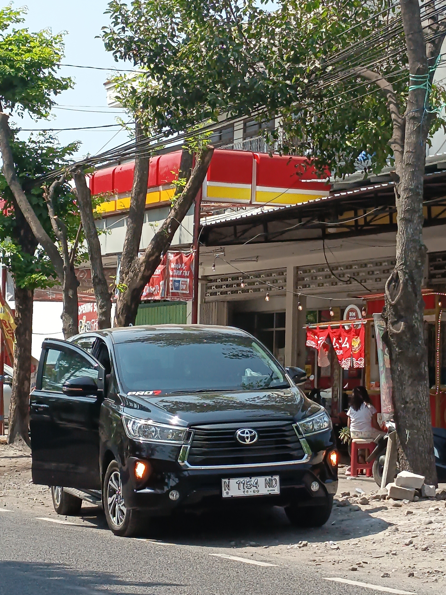 A black Toyota Innova car, license plate N 1154 ND, is parked on the side of a street in Kandangjati Kulon, Indonesia. The driver's side front door is open. The car is parked on a rough, gravelly shoulder next to a paved road. In the background, there is a building with a red and yellow awning. Several trees with green leaves line the street, their branches partially obscuring the building and the sky. A few power lines are visible strung between poles. In front of the building, there are some signs and banners, including one with red and white Japanese characters. A person in a white shirt and patterned pants is sitting on a red stool outside the building. There is also a stack of bricks near the curb and a motorcycle parked behind the person. The sun is shining, casting shadows.