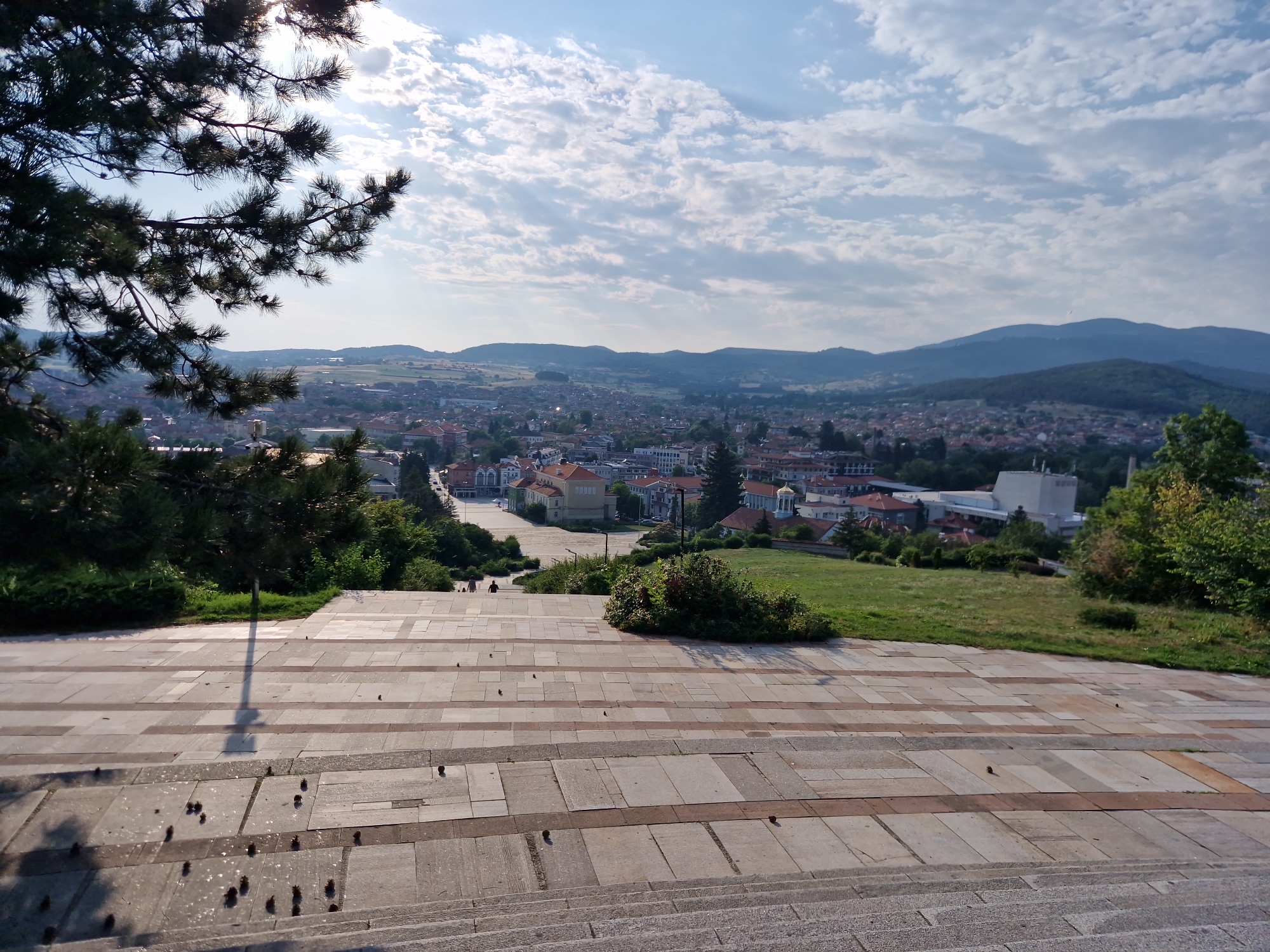 The image captures a panoramic view of Panagyurishte, Bulgaria, from a high vantage point. In the foreground, a wide staircase made of stone slabs leads down towards a plaza paved with rectangular tiles. The plaza is adorned with scattered pine cones, and the long shadow of a lamppost stretches across it.
Beyond the plaza, the city unfolds with a dense arrangement of buildings, predominantly featuring red-tiled roofs, indicative of traditional architecture. A prominent white building with a modern design stands out amongst the older structures. The cityscape is nestled within a valley, framed by rolling hills and mountains that recede into the distance. The sky is partly cloudy, with dramatic white clouds drifting across a bright blue expanse.
The greenery is lush, with mature trees and manicured grass patches evident in the mid-ground and along the slopes. Two individuals are faintly visible walking on the plaza, adding a sense of scale to the expansive scene. The overall impression is one of a peaceful, sun-drenched afternoon overlooking a charming Bulgarian town.