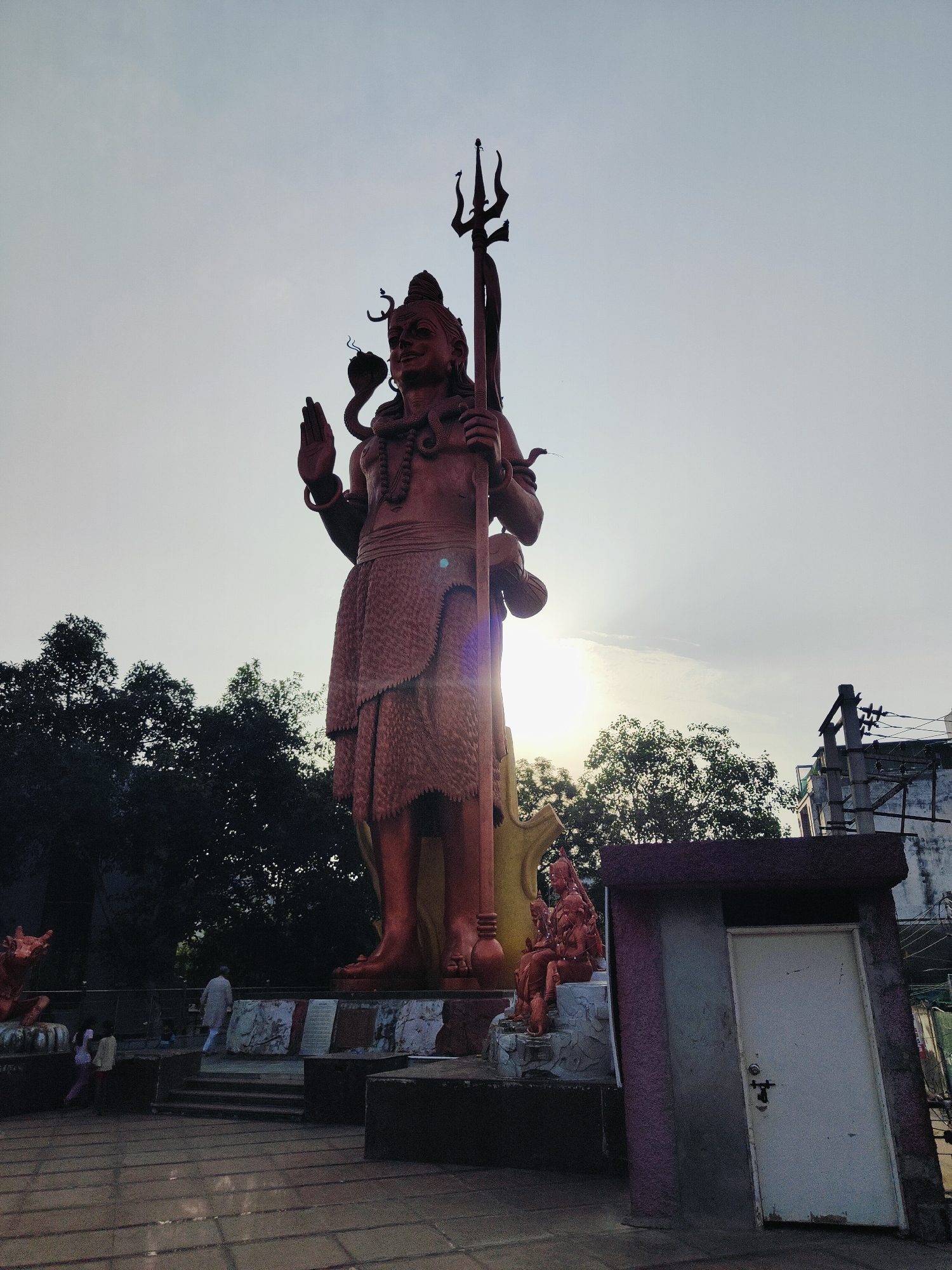 A low-angle, full shot captures a colossal statue of Lord Shiva in Gurgaon, India, bathed in the soft glow of the setting sun. The statue, rendered in a reddish-brown hue, stands tall against a pale, hazy sky. Lord Shiva is depicted in a standing posture, holding a trident in his right hand and raising his left hand in a gesture of blessing or protection. A serpent is coiled around his neck and another around his arm, both adorned with symbolic markings. He wears a skirt-like garment and elaborate jewelry.
To the right of the main statue, a smaller sculpture depicts Lord Shiva seated with another figure. In the foreground, a small, white door stands slightly ajar, set within a weathered structure. The ground is paved with square tiles, and the scene is framed by dark green trees and buildings in the background. A few individuals are visible, including a person in a light-colored robe walking up stairs toward the base of the main statue, and two children standing near the smaller sculpture. The overall atmosphere is serene and spiritual.