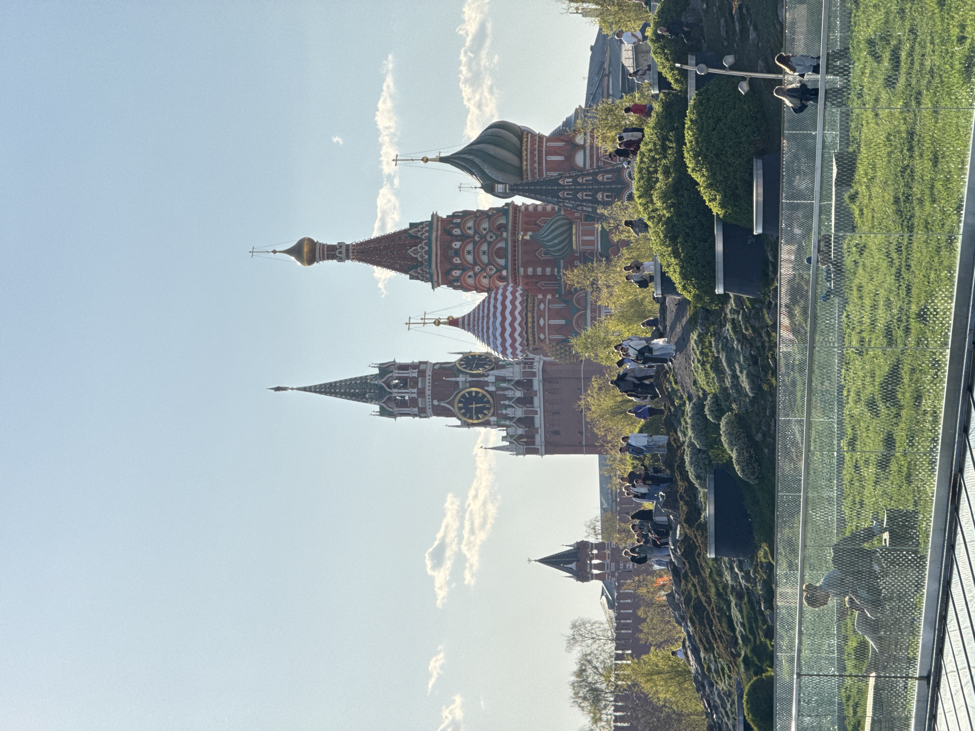 The image depicts St. Basil's Cathedral and the Spasskaya Tower of the Moscow Kremlin in Moscow, Russia, under a clear sky with scattered clouds. A glass walkway extends into the foreground with people walking on it. Trees and shrubs line the walkway. People are sitting and standing along the hillside overlooking the historical buildings.