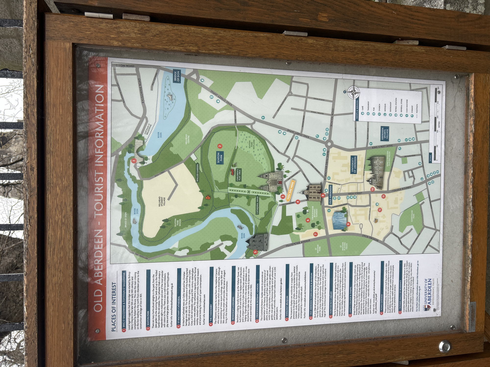 The image shows a wooden-framed tourist information board displaying a map of Old Aberdeen, a historical part of Aberdeen City, United Kingdom. The map outlines key points of interest, including the Brig o' Balgownie, Seaton Park, St Machar's Cathedral, the Chanonry, Benholm's Lodging, Cruickshank Botanic Garden, the Zoology Museum, the Old Aberdeen Town House & Museum, the Duncan Rice Library, King's College & Chapel, the Powis Gateway, and Crombie Halls of Residence. The map also indicates the locations of various shops and services such as cafes, takeaways, pubs, churches, mosques, bookshops, petrol stations, general stores, cash machines, and chemists. The information board features text descriptions for each point of interest, providing historical context and details. The River Don winds through the map, and the University of Aberdeen logo is displayed at the bottom. The environment around the board includes a gray metallic object.