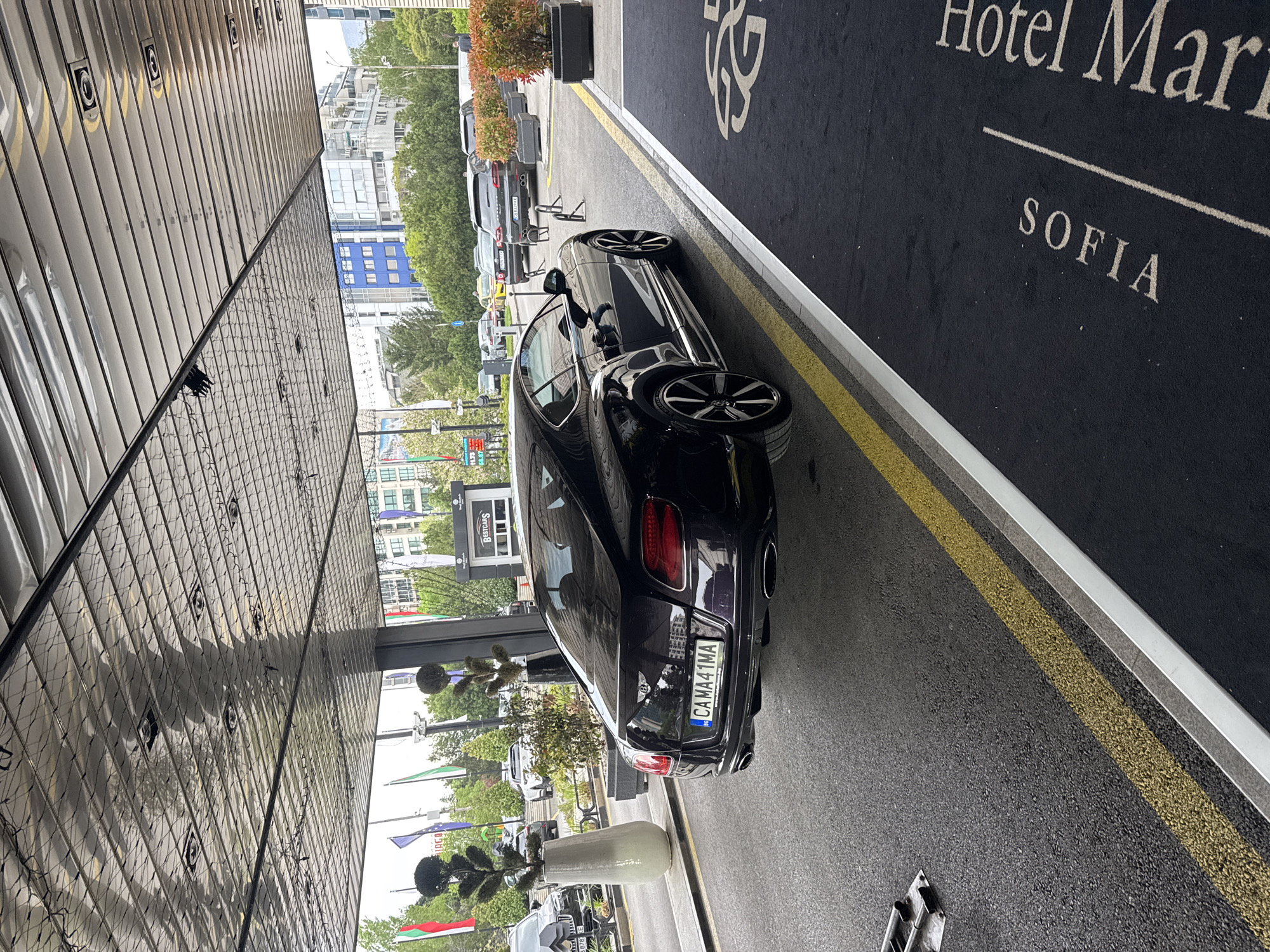 The image captures a black luxury car, appearing to be a Bentley, parked at the entrance of a hotel in Sofia, Bulgaria, during daytime. The vehicle is positioned on an asphalt driveway bordered by a yellow line and a silver curb. Adjacent to the curb is a large black outdoor mat or carpet, which displays "Hotel Marri" (likely referring to Marriott) and "SOFIA" in light-colored text, confirming the city.
The rear of the car faces away from the viewer, showing its license plate "CAMA41MA" with the "BG" country code. The building's facade is modern, featuring large reflective glass panels that show distorted reflections of the surroundings. Potted plants and ornamental shrubs are arranged along the driveway and near the entrance.
In the background, other vehicles are visible, parked along a street. A sign for "Bestcars" can also be seen further down the road. Several flags are fluttering in the distance, including the Bulgarian flag with its distinctive white, green, and red horizontal stripes. The weather appears bright, possibly overcast, as suggested by the even lighting and lack of harsh shadows. There are no prominent human figures or specific activities taking place, other than the car being present at the hotel entrance.