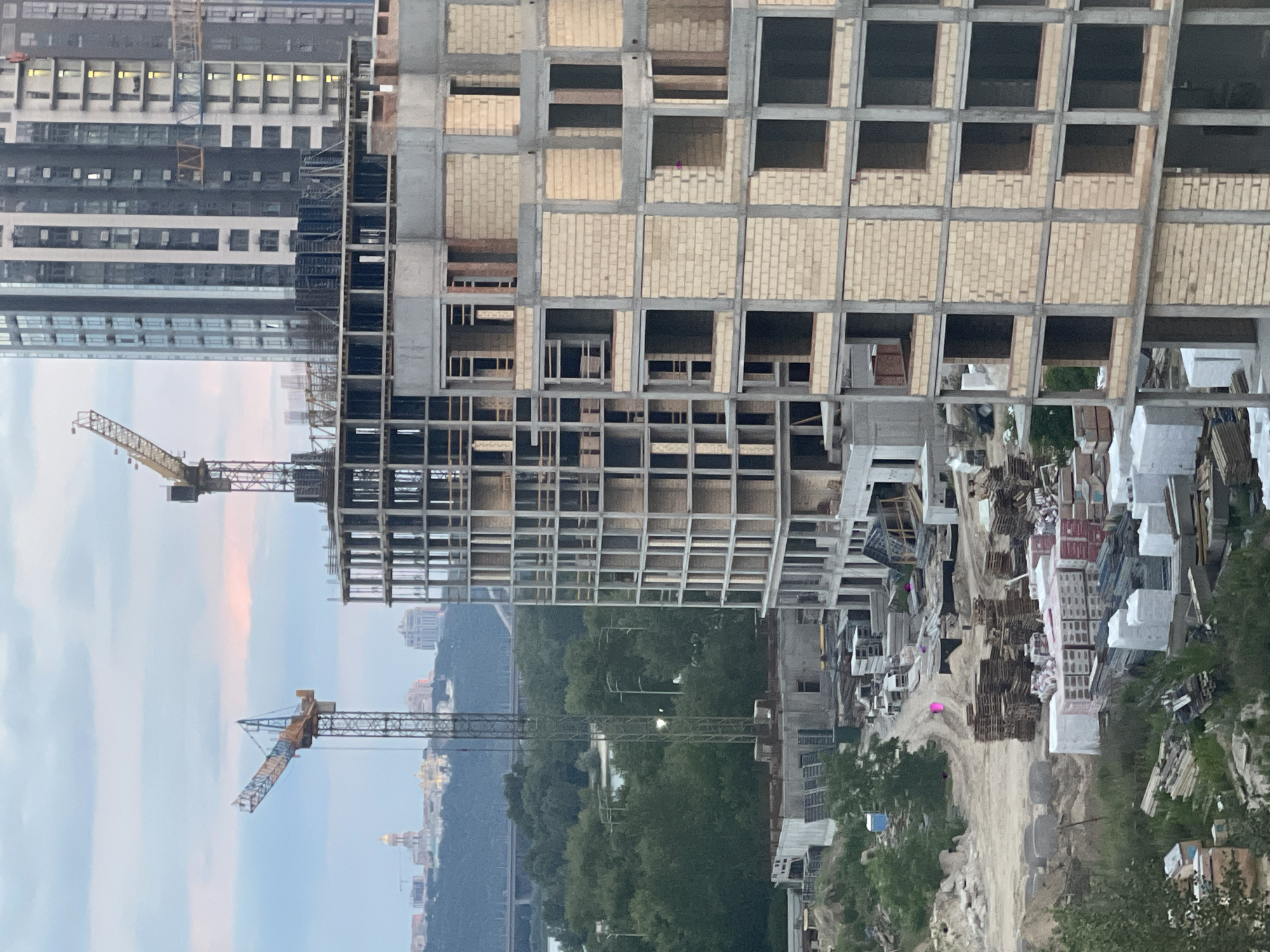 This image depicts an ongoing construction site in Kyiv, Ukraine, characterized by multiple tall buildings in various stages of development. In the foreground, a partially constructed concrete and brick building dominates the frame, showcasing its skeletal structure with visible floors, columns, and window openings. Adjacent to it, another building, further along in its construction, is a taller, modern high-rise with a dark facade and numerous windows.
Two prominent construction cranes are visible. One, a large yellow tower crane, stands tall beside the skeletal building, suggesting active work. Another, a smaller crane, is positioned in the background, partially obscured by the taller structures.
The surrounding environment includes a scattering of construction materials such as stacks of pallets, bags of building supplies, and piles of sand. Green trees and vegetation line the mid-ground, with a body of water and distant cityscape visible beyond. The sky is overcast with a hint of pink, indicating either dawn or dusk. The overall scene conveys a sense of urban development and progress.