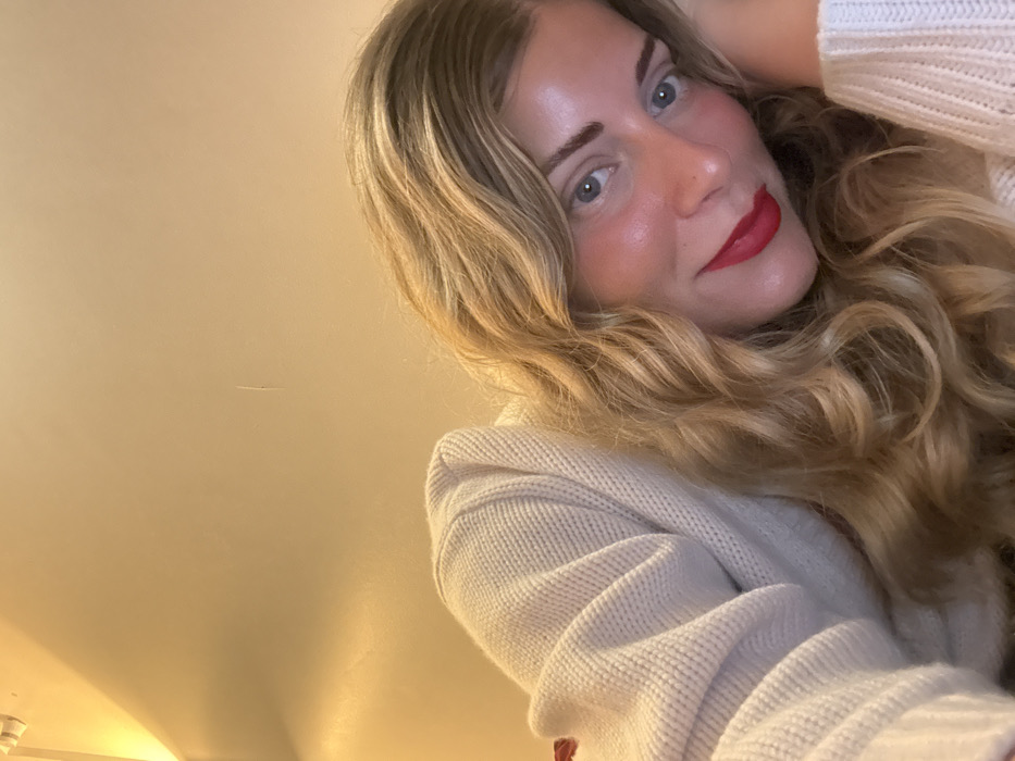 The image displays a close-up, diagonal shot of a woman in an indoor setting. She possesses long, wavy blonde hair, light-colored eyes, and fair skin, accentuated by red lipstick and blush. She is wearing a light-toned, possibly off-white, ribbed knit sweater. Her head is tilted, resting on her left arm, which is covered by the sweater sleeve. She gazes directly at the camera. The background is a plain, warm beige wall, illuminated by an overhead light fixture visible in the upper left corner. A faint vertical mark is present on the wall towards the upper right. No identifiable features within the image indicate its location in Paris, France.