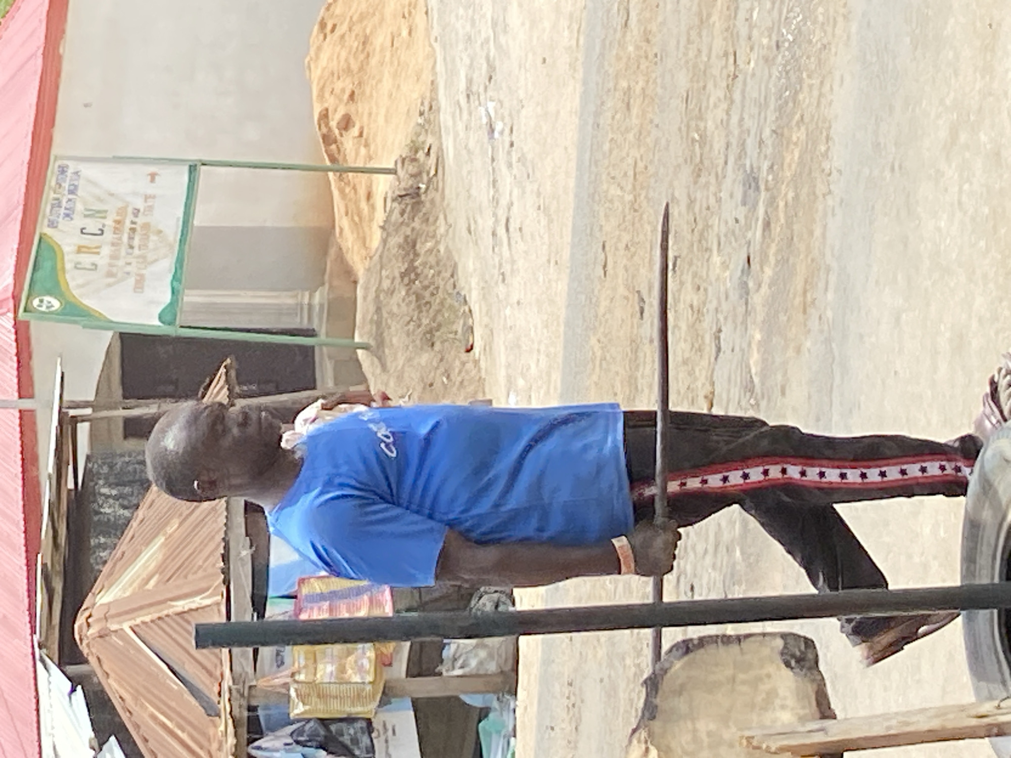 This image, taken in Mararaba, Nigeria, captures an outdoor scene during daylight hours, characterized by bright, even lighting and a dry, dusty environment.
The central subject is a man of dark complexion, standing upright (the image is rotated 90 degrees clockwise). He is bald and wears a bright blue short-sleeved t-shirt with some white text, and dark trousers adorned with a distinctive red and white star-patterned stripe running down the side. An orange wristband is visible on his left arm. He is holding a long, slender, dark metal rod horizontally with both hands, suggesting he is actively engaged in work or a specific task. His expression appears neutral and focused.
In the background, behind and to the left of the man, are rustic structures made of wood and corrugated metal, possibly forming a stall or shed. A large vehicle tire is partially visible at the bottom right corner of the frame. The ground is bare earth or sand.
Behind the man, a light-colored building wall is visible, in front of which stands a green and white sign. The sign prominently displays "CRCN" in large letters. Other readable text on the sign, though partially obscured or difficult to decipher, includes "REGIONAL HEADQUARTERS," "DUAVU NIGERIA," "EKOMU NDUAK," "CONGREGATIONAL," and "CROSS RIVER STATE." There's also a line that seems to read "2018 CEASED IN 1952" or similar. The setting suggests a community or commercial area, likely a market or roadside in a semi-urban or rural part of Nigeria.