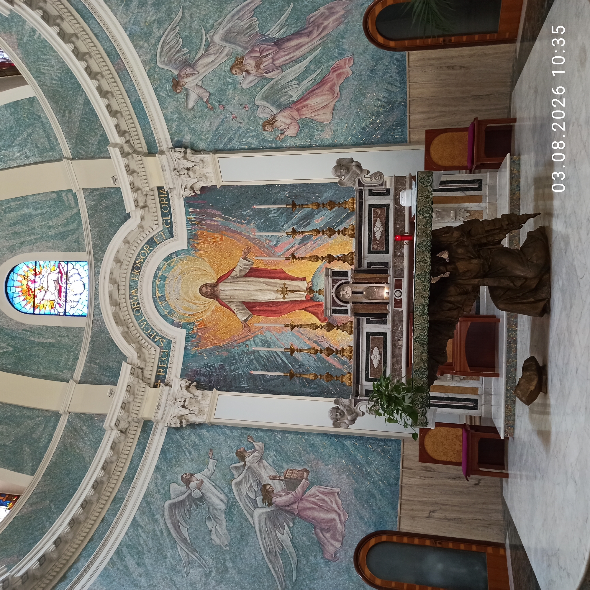 The image depicts the interior of a church in Ruvo di Puglia, Italy, specifically the apse area. Dominating the scene is a large mosaic mural of Jesus Christ with his arms outstretched, radiating a golden light. The mosaic is flanked by depictions of angels on the walls. In the foreground, a marble altar is supported by a sculpted kneeling figure. Several candelabras and candles are present on the altar. To the left and right of the altar are wooden chairs with upholstered seats. The floor is made of white marble. The overall environment is richly decorated, suggesting a place of worship. A date and time stamp "03.08.2026 10:35" is visible in the lower right corner.