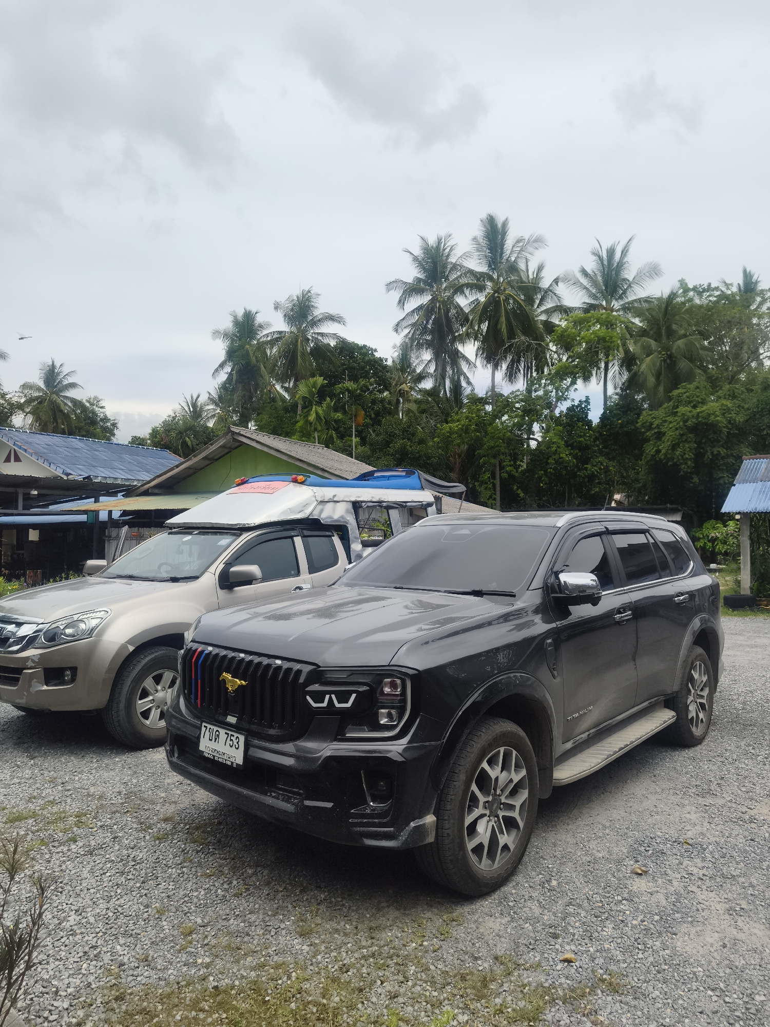 The image shows two SUVs parked on a gravel lot. The SUV in the foreground is black and has a modified grille with red, white, and blue accents and a gold horse emblem. It also has a Thai license plate that reads "7 บฉ 753". The SUV in the background is gold and appears to be a pickup truck with a covered bed. Behind the vehicles are several buildings with corrugated metal roofs, palm trees, and lush green vegetation. The sky is overcast. There are no people visible in the image.