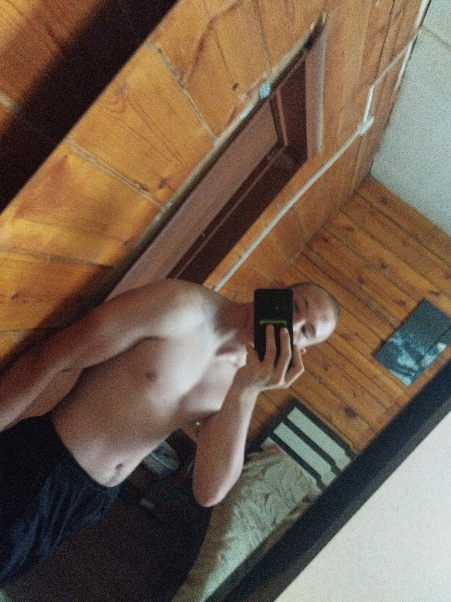 The image shows a shirtless individual wearing black shorts, taking a mirror selfie. The individual is in a room with wooden paneling on the walls and ceiling. A mirror is visible, reflecting the individual and the room. In the reflection, a bed with light-colored bedding is visible. There is also a small dark rectangular object on the wall in the reflection, possibly a picture or a decorative item. The lighting appears to be natural, coming from an unseen source. The room appears to be a bedroom or a similar personal living space. The image itself is slightly blurry and grainy. The location is identified as the city of Kirensk, Russia.