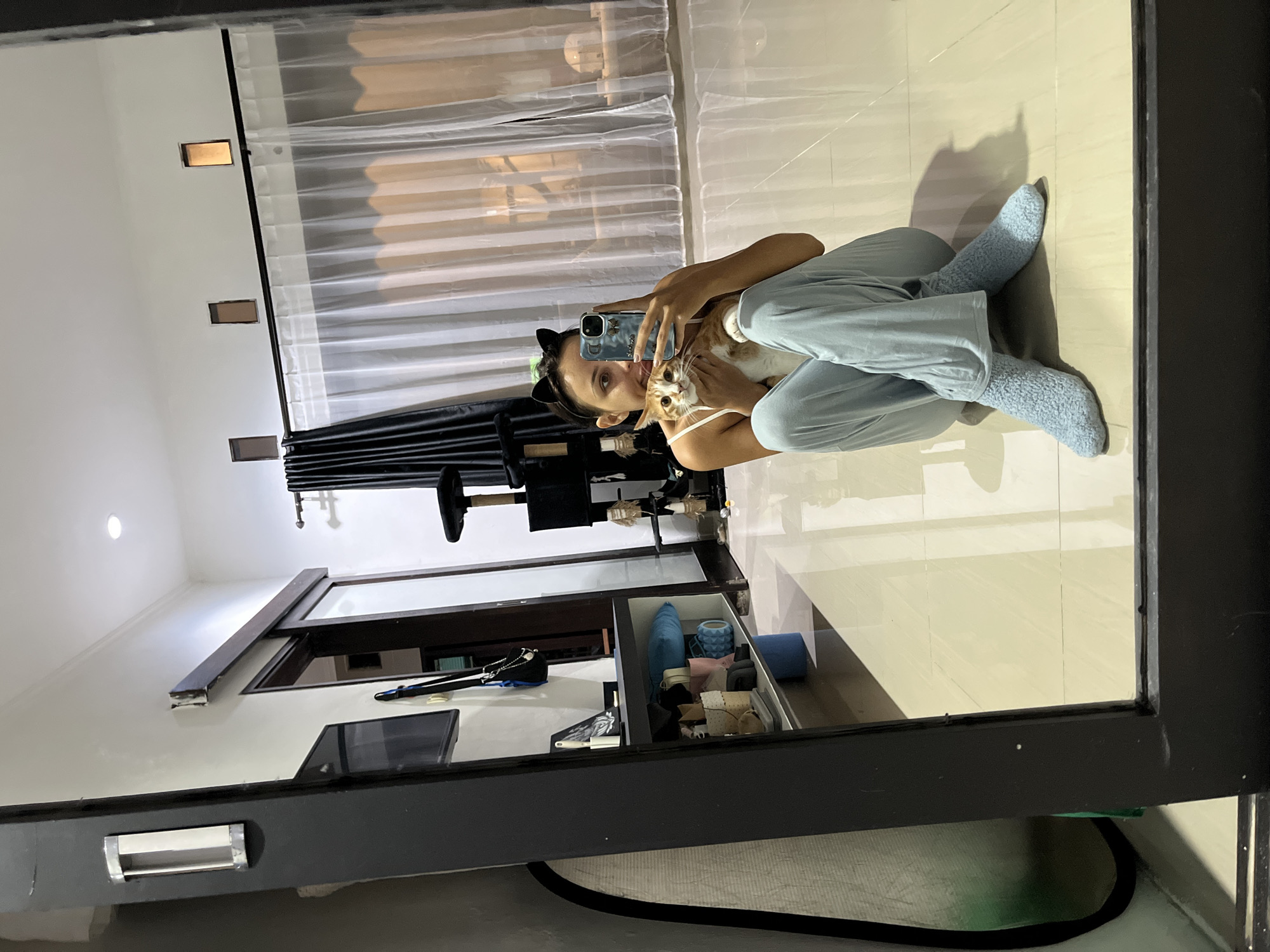 A woman wearing cat-ear headwear and blue pajama pants and socks is sitting on a tan-tiled floor, holding a cat and taking a selfie with a cell phone. The reflection shows a room with white walls, a sheer curtain, a dark curtain, a cat tree, and a storage unit. The location is likely a private residence, but more details are needed for specific identification within Kuta Selatan, Indonesia.