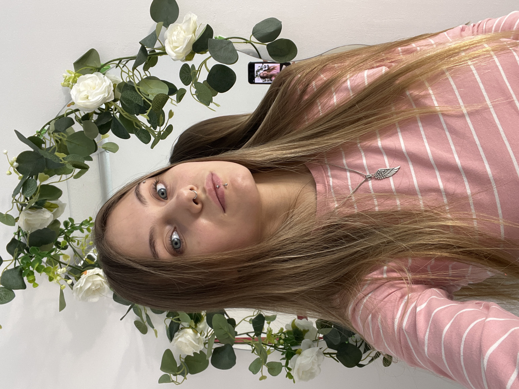 The scene captures a female taking a mirror selfie in an indoor setting in Foki, Russia. The subject, positioned centrally and looking directly at the camera through the mirror, has long, straight, light brown hair with blonde highlights, blue eyes, and a fair complexion. She is wearing a pink long-sleeved shirt with thin white horizontal stripes and a silver necklace featuring a winged pendant. She has a silver ring piercing in her left nostril, a silver stud piercing above her left eyebrow, and a silver bar piercing in her philtrum area. The mirror is partially framed by an artificial floral garland consisting of white roses and green leaves, against a plain white wall. A black smartphone, held by the subject's right hand, is visible in the reflection, displaying the subject's image on its screen.