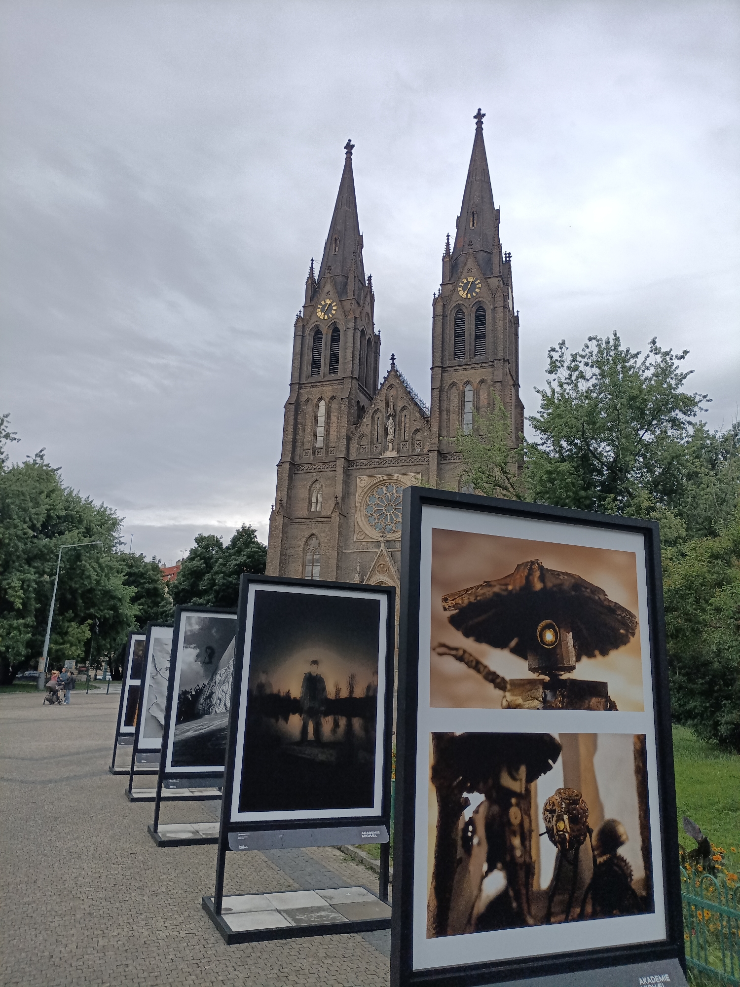This media file shows an outdoor exhibition of photographs in front of a gothic-style church with two prominent spires and a clock face on one. The sky is overcast and cloudy, suggesting a daytime setting with potentially gloomy weather. The exhibition consists of several large, framed photographs displayed on stands along a paved walkway.
The photographs themselves appear to be a mix of black and white and color images. Some of the black and white photos feature dramatic lighting and potentially dark or somber themes, including a silhouette of a person standing by a body of water and another with graffiti. The color photographs in the foreground are close-ups of what appear to be intricate, possibly metallic or mechanical sculptures or objects, some with a steampunk aesthetic.
In the background, to the left of the exhibition, there are trees and a few people walking, one pushing a stroller, suggesting a public space like a park or plaza. The architecture of the church, with its pointed arches, stained glass windows, and ornate stonework, is a significant element of the scene. The overall impression is that of an art installation set within a historical and architectural context.
Visible text includes "AKADEMIE" on the bottom right corner of the frame, suggesting the exhibition might be related to an academy or educational institution.