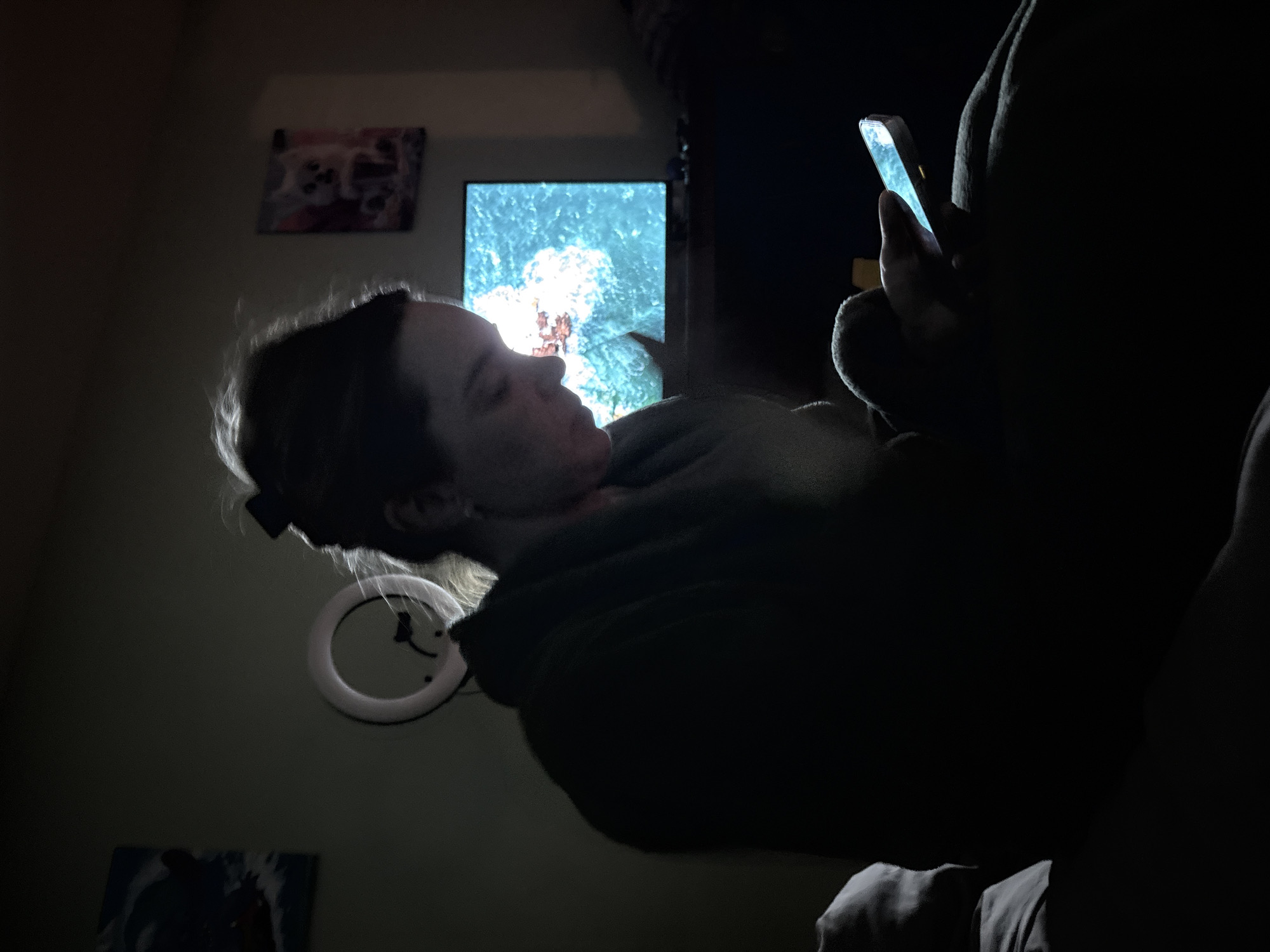 A woman is lying in a darkened room, illuminated by the screen of a mobile phone she holds in her hand. Behind her, on the wall, is a flat-screen TV displaying an image of what appears to be marine life or an underwater scene. There's also a circular light fixture, possibly a ring light, mounted on the wall. Additionally, there are framed pictures visible on the wall, one to the left of the TV and another below. The lighting suggests it is nighttime. The image does not contain information to prove that this image was taken in the city of Sinyavino, Russia.