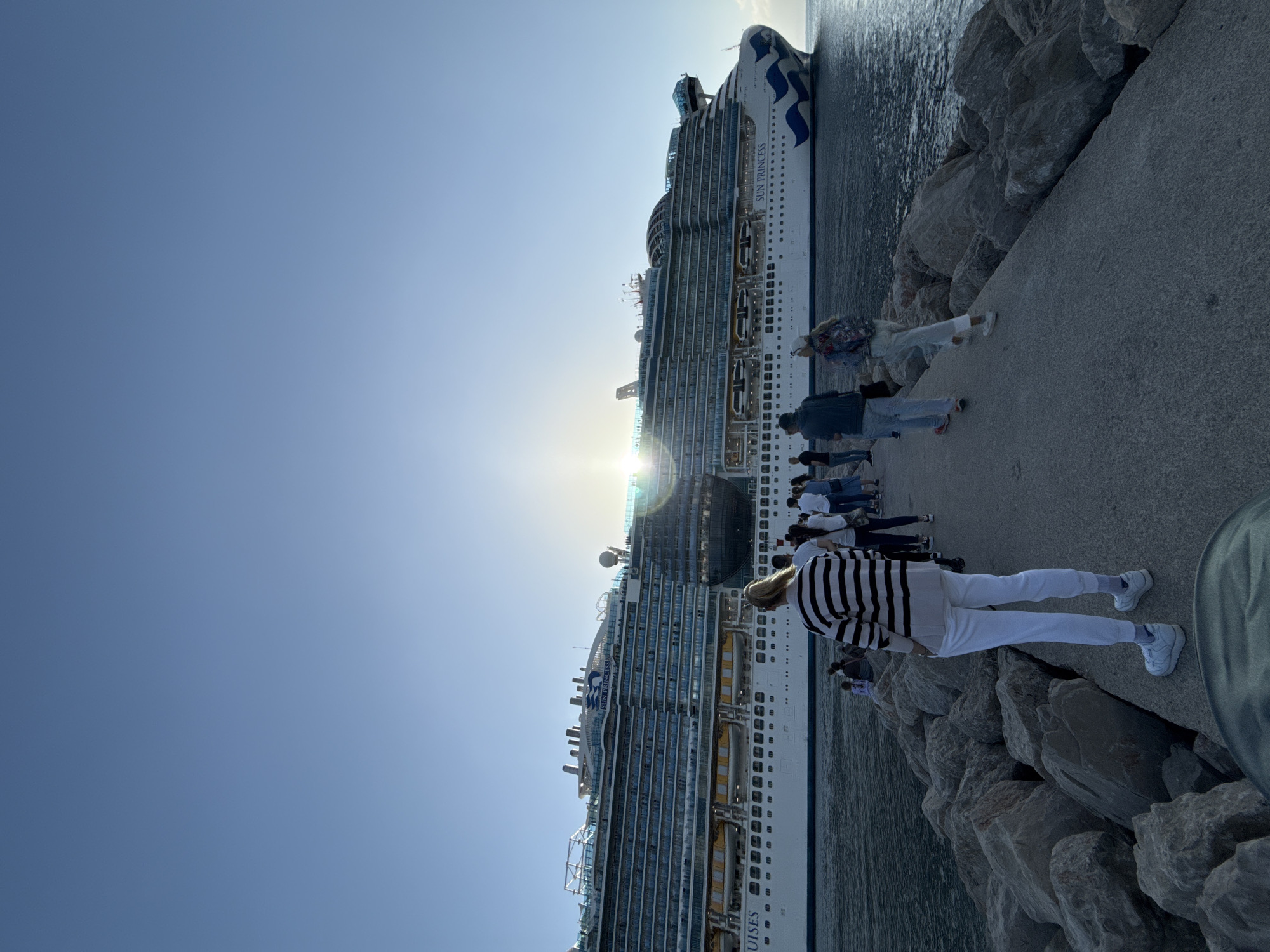 The image shows two large cruise ships docked in Bar, Montenegro. A crowd of people is standing on a concrete pier alongside the ships, with large rocks forming a barrier between the pier and the water. The sky is clear and blue, and the sun is positioned between the ships, creating a glare. One of the ships is partially identified as the "Sun Princess." The overall environment suggests a sunny day at a port. People are likely disembarking or embarking on the cruise ships.