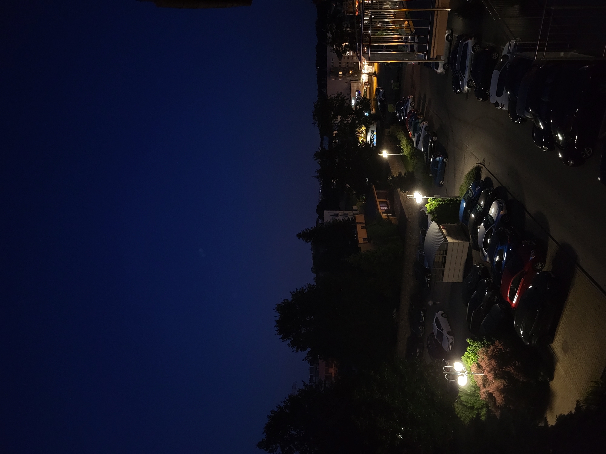 A nighttime aerial shot shows a parking lot filled with cars and some trees. The sky is dark blue, indicating it is either dusk or night. Streetlights illuminate parts of the parking lot, casting long shadows. A few buildings and trees are visible in the background, their details obscured by the darkness. The image has a high contrast due to the artificial lighting against the dark environment.