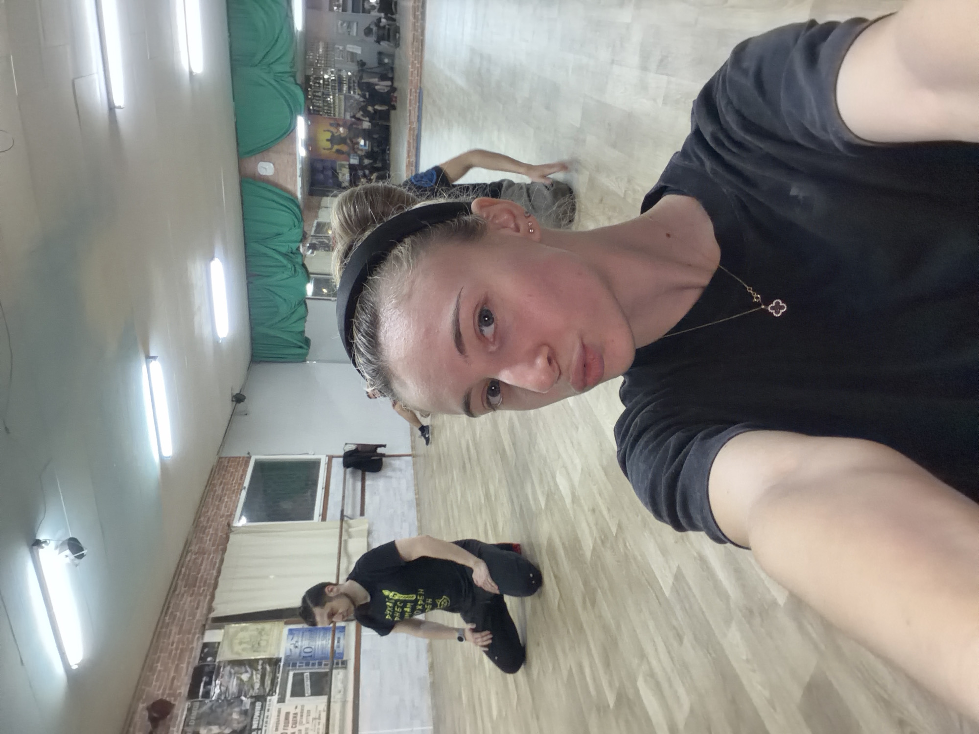 A selfie taken in a dance studio in Plovdiv, Bulgaria, shows a person with fair skin, wearing a black t-shirt and a black headband, positioned in the foreground. A gold necklace with a clover-shaped pendant is visible on their chest.
In the background, there are two other individuals performing dance movements on a light-colored wooden floor. One is a man wearing a black t-shirt and black pants, kneeling on the floor. The other, further away, is kneeling as well.
The dance studio has green curtains along one wall and several windows. The walls are decorated with posters and notices. Fluorescent lights illuminate the room.