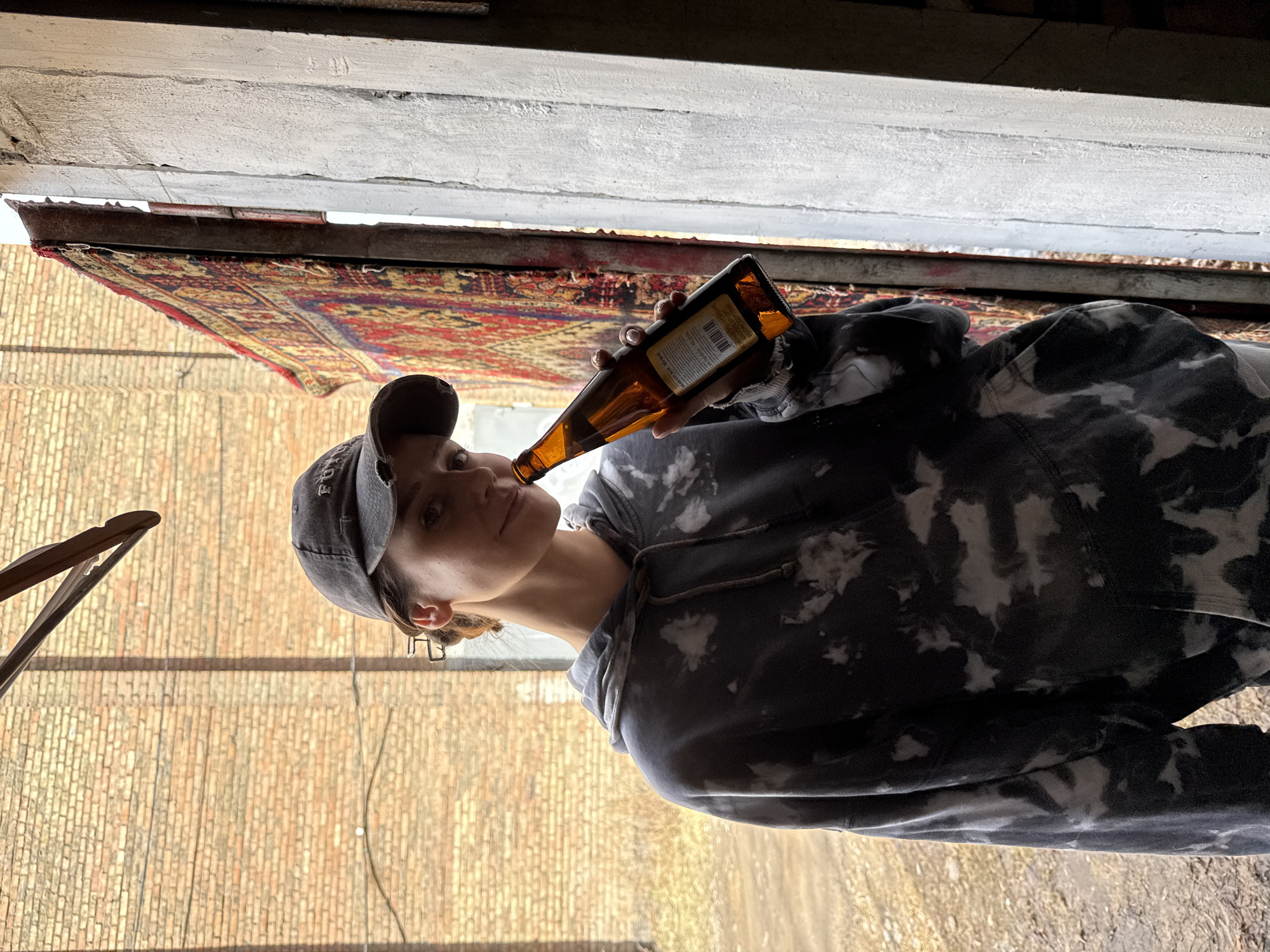 A woman with a cap is drinking from a brown beer bottle. She is wearing a black and white patterned hoodie and glasses. She is standing outside in a space with a brick wall. An ornate rug is hanging overhead. The location appears to be Krasnoyarsk, Russia.