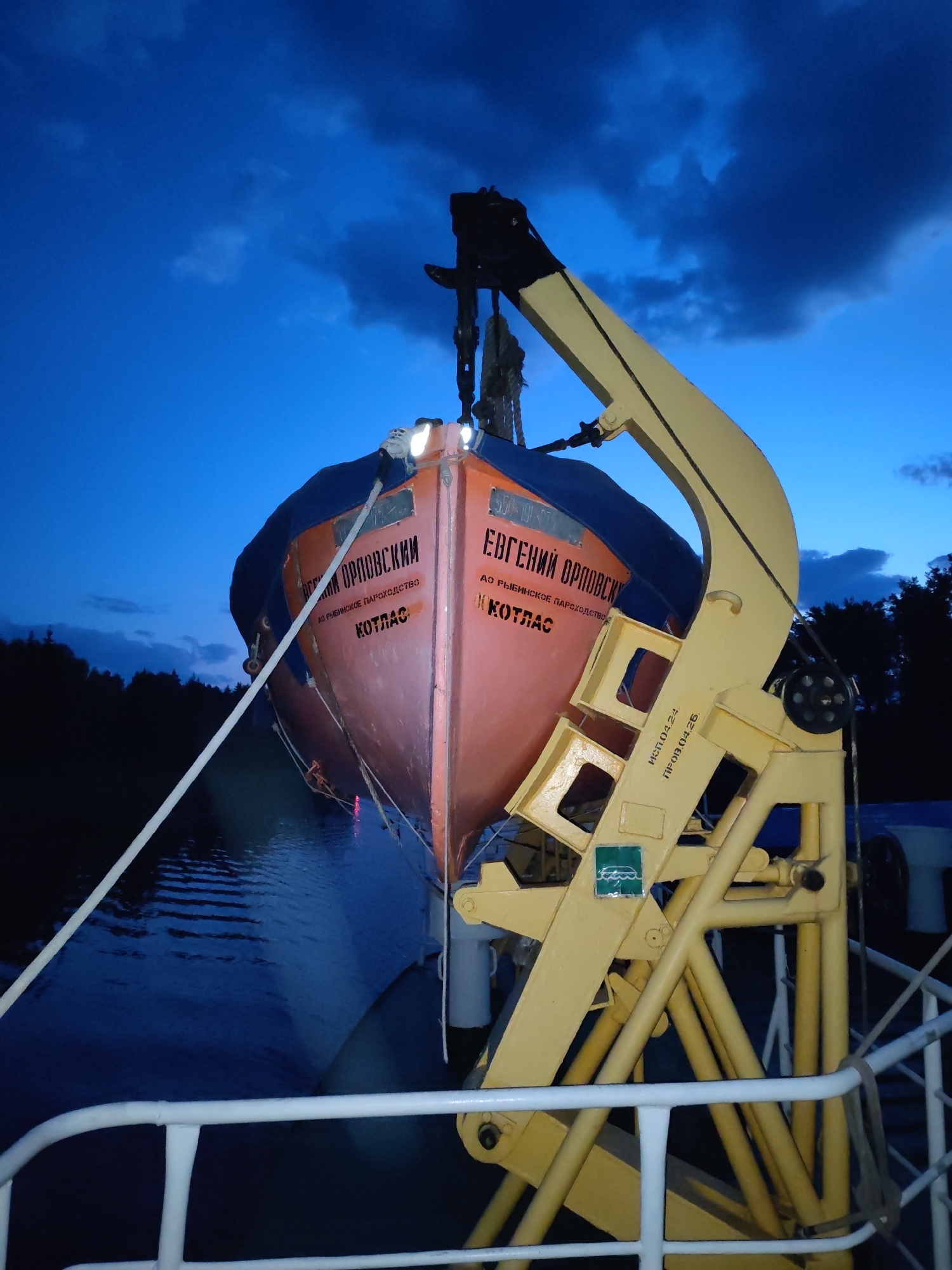 The image shows a lifeboat, painted orange and blue, suspended by a yellow davit system on a ship. The lifeboat has text written in Cyrillic on both sides: "ЕВГЕНИЙ ОРЛОВСКИЙ AO РЫБИНСКОЕ ПАРОХОДСТВО КOTЛАС" (Yevgeniy Orlovskiy, Rybinsk Shipping Company, Kotlas). A white rope is attached to the front of the lifeboat. The davit system has a green sticker with a symbol on it and more text in Cyrillic that is partially visible: "ИСП.04.24. ПРОВ.04.26." (ISP.04.24. PROV.04.26.). The photo appears to be taken at night or dusk, with a dark blue sky and clouds visible in the background. The trees on the shore are visible in the distance. The surface of the water and the ship's railing are also visible. The image does not specify the exact location within the city of Unknown, Unknown.