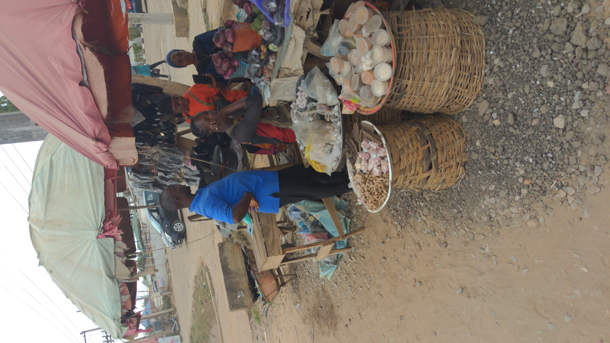 This image captures a vibrant outdoor market scene in Ado, Nigeria, during daytime. The setting is characterized by unpaved ground, a mix of packed dirt and gravel, where multiple rustic wooden stalls are set up. Large canopies, one pink and another light green/white, provide essential shade over the market area.
Several people are visible, indicating ongoing market activity. In the immediate foreground, a man dressed in a blue t-shirt and dark trousers stands near one of the wooden stalls. Behind the stalls, various women are present, seemingly engaged as vendors or shoppers. One woman with a head covering is seen next to a large woven basket filled with red and purple onions. Another woman, wearing an orange top, appears to be sitting or stooping, holding a mobile phone. A third woman with dark skin is also seated behind a display of goods.
The stalls are laden with a variety of produce and items. Prominently displayed in large, woven baskets are diverse goods: one basket holds a generous amount of onions, another contains numerous small, clear plastic containers filled with a light-colored substance (possibly spices or grains), and a third basket is neatly arranged with garlic bulbs and ginger roots. Other goods, including packaged items and what might be dried fish, are laid out on wooden tables. In the background, a black car is partially visible, parked on what appears to be a road adjacent to the market. The diffused natural light suggests it's either morning or afternoon. No specific emotions are distinctly visible on the faces of the individuals, and no discernible text can be read in the image. The scene portrays a typical, bustling local market in action.