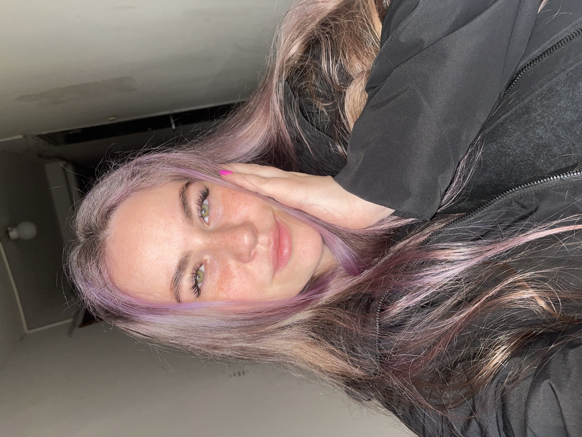 A selfie-style portrait of a young woman with dyed hair. Her hair is primarily brown with a lavender shade around the face. She has fair skin, green eyes, and freckles. She is wearing a black jacket and her hand is placed against her cheek. The background appears to be a dimly lit interior space with a white ceiling. The location in the city of Chaikovsky, Russia is not directly visible in the image.