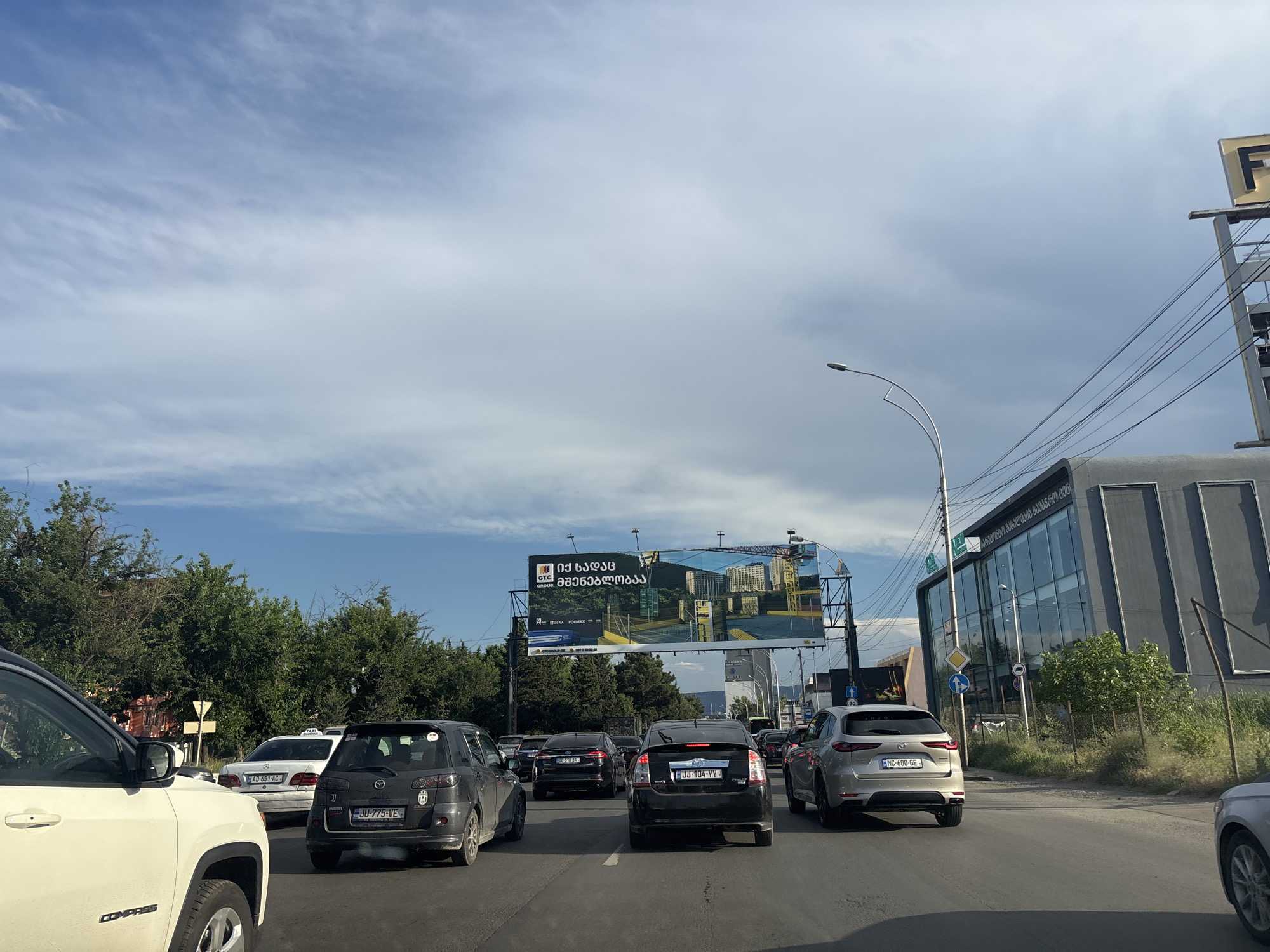 The image depicts a traffic jam on a multi-lane road in Tbilisi, Georgia, under a partly cloudy sky. Cars are stopped in various lanes, with vehicles visible including a white Jeep Compass in the foreground, a black Mazda, a silver SUV, and other cars. A billboard advertising real estate development, featuring a crane and buildings, is prominently displayed above the roadway. Adjacent to the billboard is a modern, grey building with large glass windows. Streetlights and power lines are also visible, indicating an urban environment. The presence of traffic and signage suggests a bustling city scene.