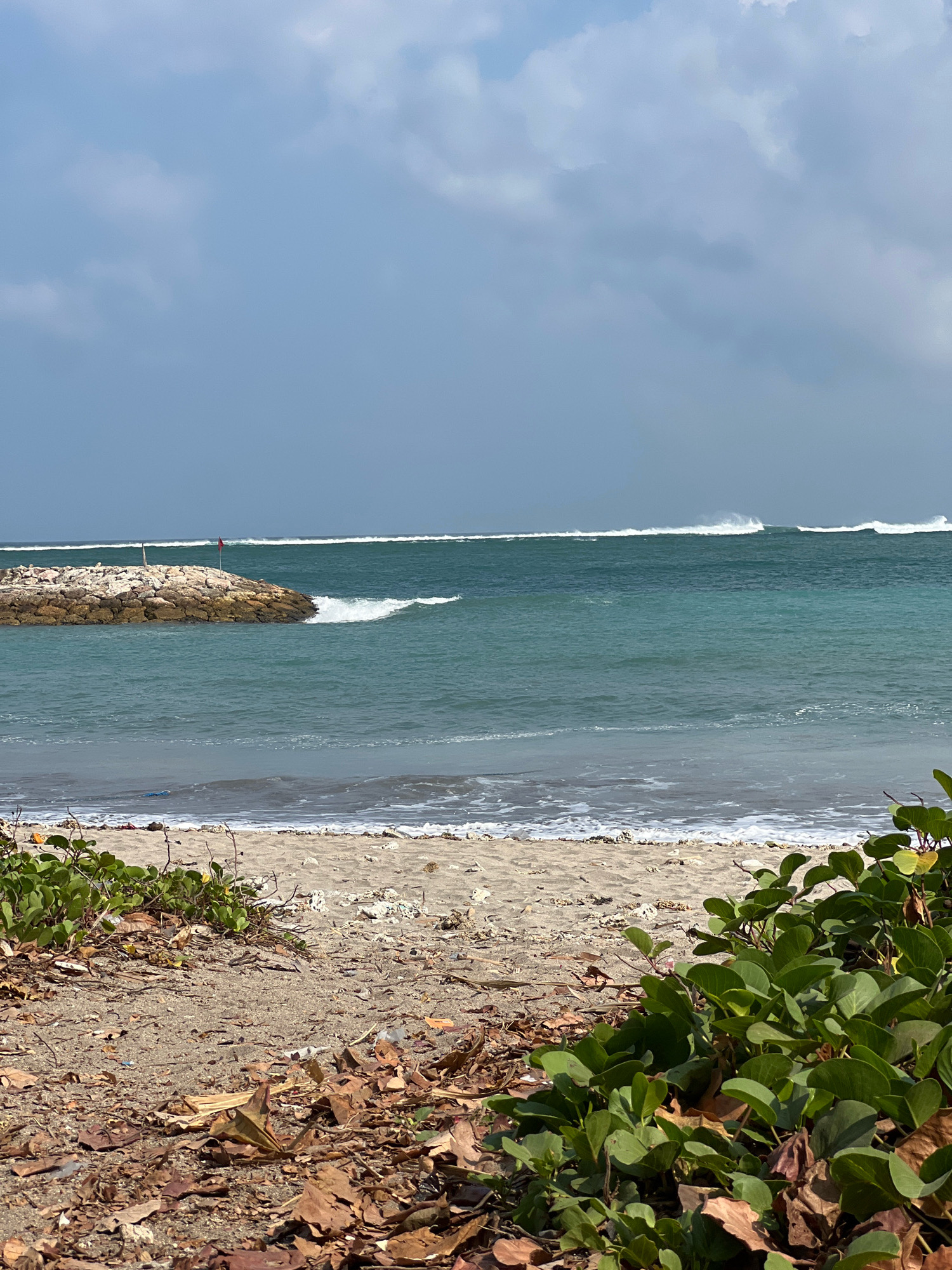 A beach scene in Kuta, Indonesia, under a partly cloudy sky. The foreground is dominated by a sandy beach covered with fallen brown leaves and some green vegetation. The middle ground features the ocean with turquoise water and waves breaking. A stone jetty, adorned with a small red flag, juts out into the water on the left side of the frame. In the background, the ocean stretches to the horizon, with more waves visible. The overall impression is of a natural, coastal environment.