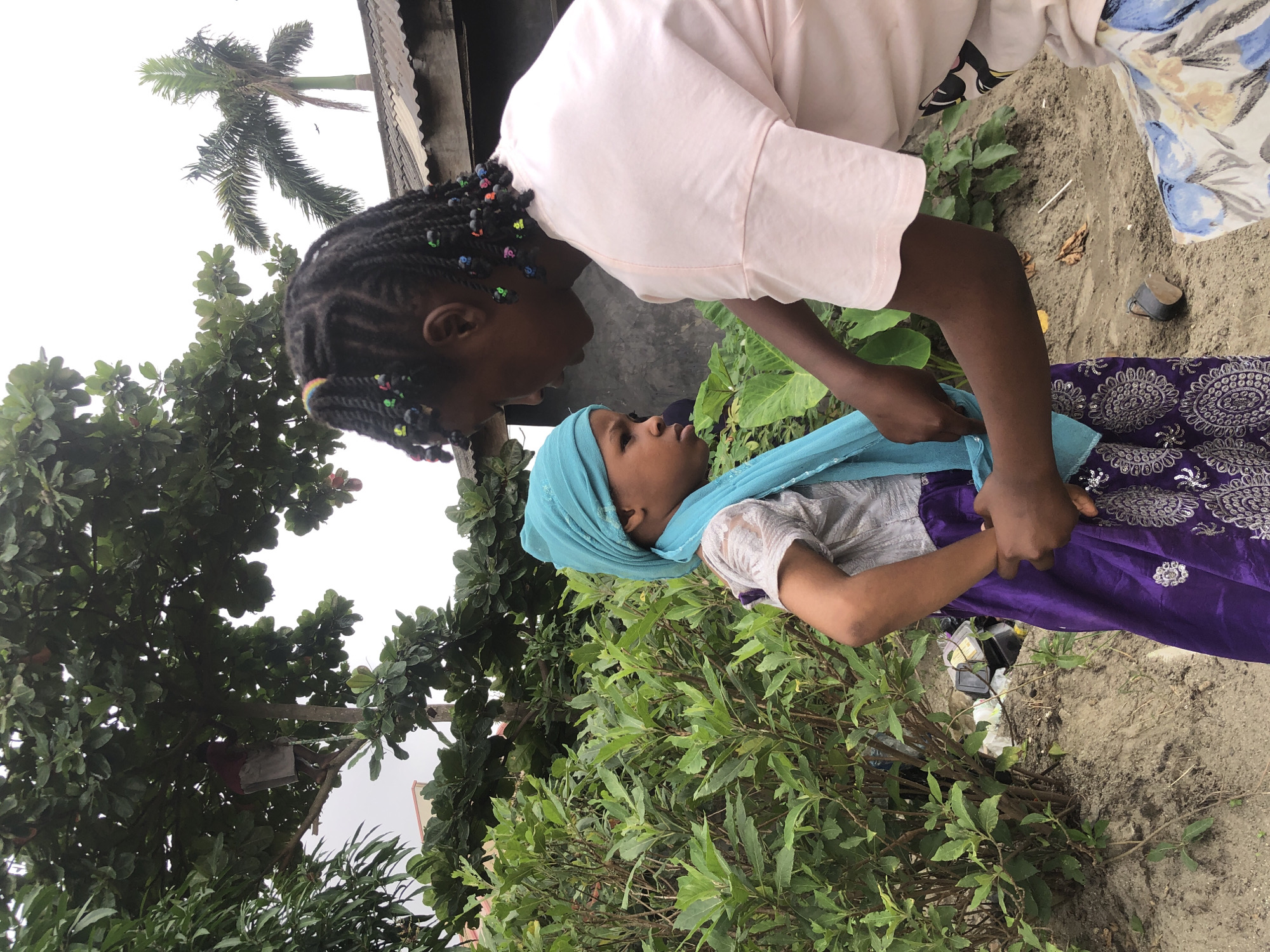 Two young Black females stand in an outdoor environment. The female on the left wears a light gray shirt and a purple and silver patterned skirt, along with a turquoise headscarf. The female on the right wears a light pink t-shirt and a floral skirt, and her dark hair is styled in braids adorned with colorful beads. The female on the right appears to be adjusting the headscarf of the female on the left. They are positioned in front of lush green foliage and a sandy embankment. In the background, a tin-roofed structure and a palm tree are visible, suggesting a tropical or subtropical urban setting. The overall scene suggests a candid moment captured outdoors, possibly in a residential or informal area.