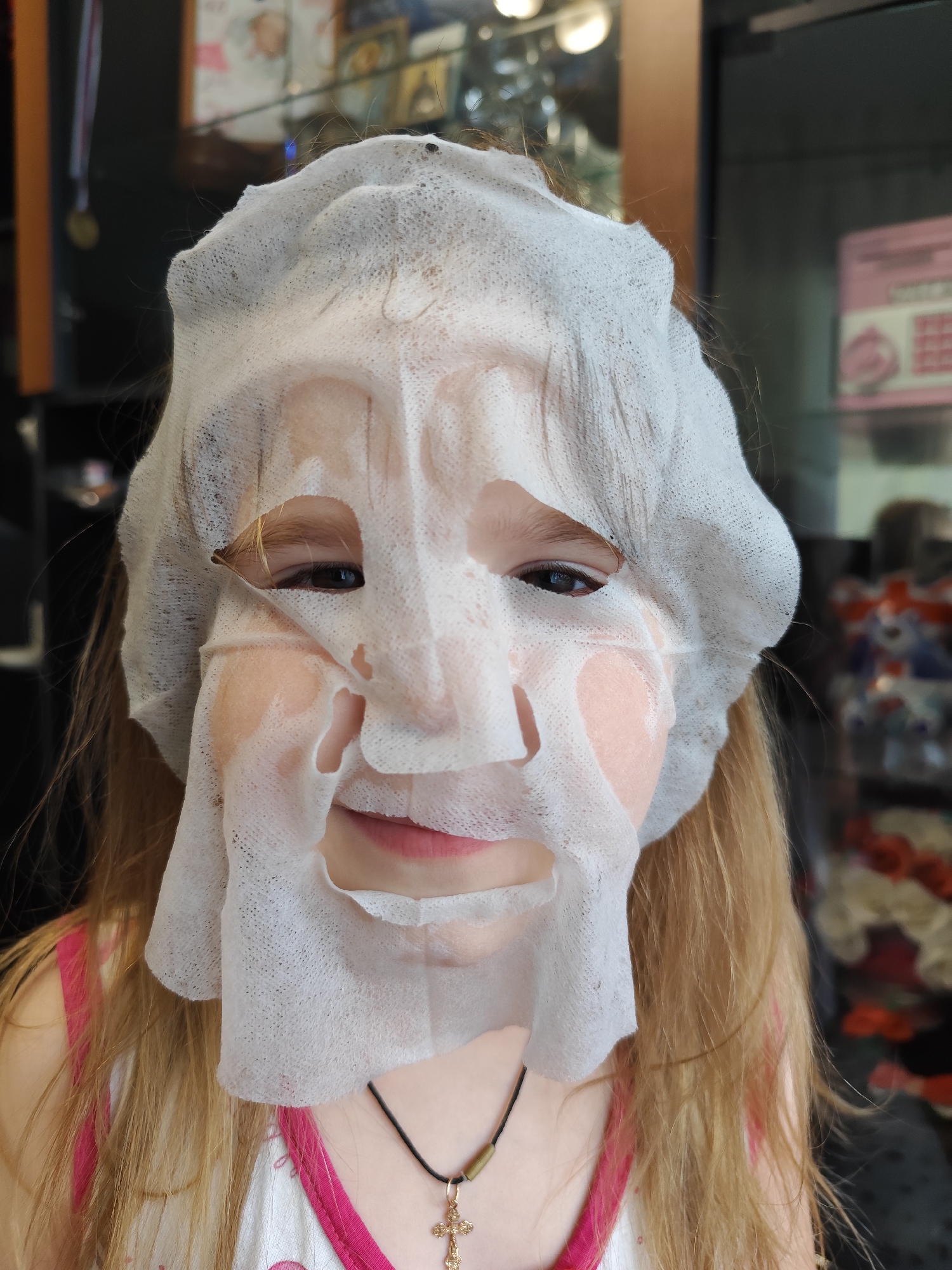 A young, fair-skinned child with long blonde hair is wearing a white sheet mask on their face, with the mask covering most of the face except for the eyes and mouth. The child is wearing a pink-strapped shirt and a necklace with a gold cross pendant. The background is blurry but appears to be a shop or store interior. The location in the city of Фоки, Russia is not determinable from the image.