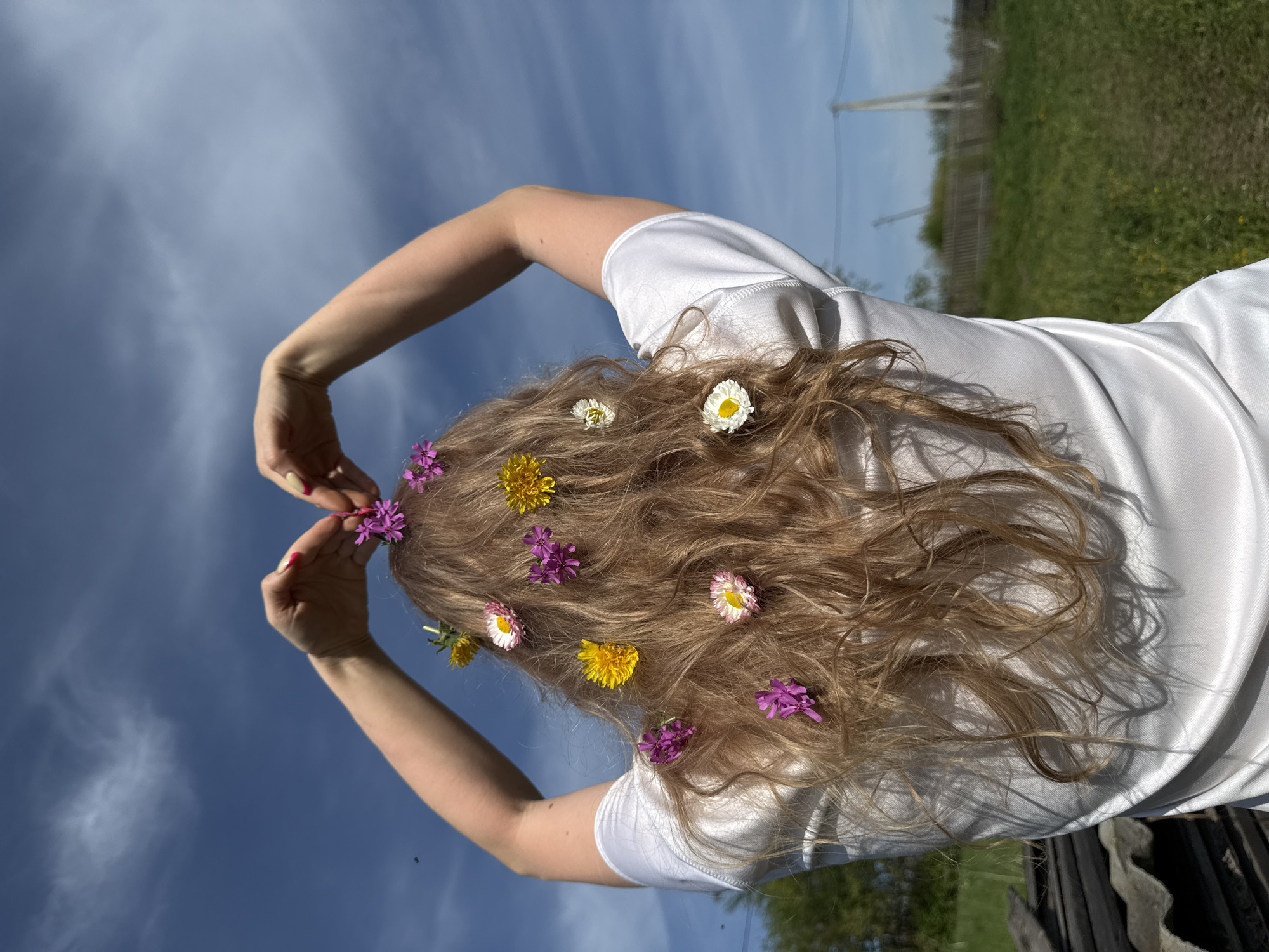 A woman with long, wavy blonde hair is facing away from the viewer, outdoors on a bright day, with a blue sky dotted with scattered clouds. She is wearing a white t-shirt and is in the process of placing small, colorful flowers in her hair. Her arms are raised, and her hands are positioned near the top of her head as she adds the flowers, which include purple, yellow, and white varieties. The background shows a blurred landscape with green vegetation and a hint of a wooden fence. The image is taken from a low angle, emphasizing the sky and the woman's posture. The location is Nizhnyaya Chermoda, Russia.