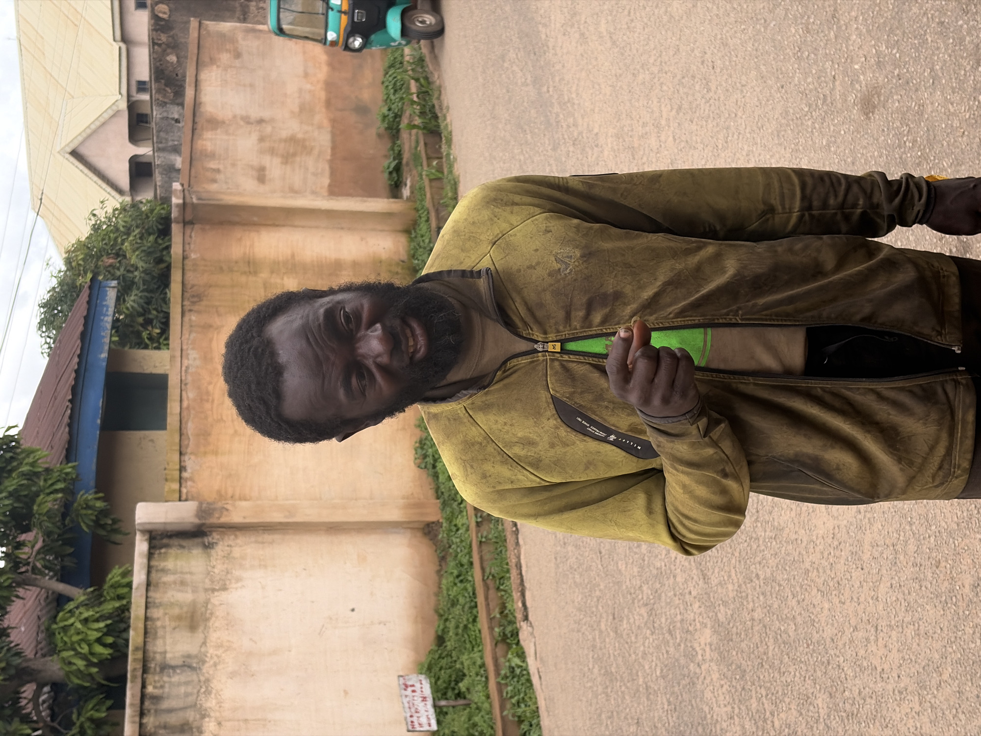 A man with a beard and dark skin, wearing a green t-shirt and a worn, olive-green jacket, is pictured in what appears to be an outdoor, urban setting in Jos, Nigeria. He is looking towards the camera and holding up his right index finger, possibly gesturing. Behind him, a building with beige walls and a blue and red-striped roof is visible, along with some green foliage. Further in the background, another building with a light-colored, pitched roof can be seen. A turquoise auto-rickshaw is parked on the right side of the frame, on a paved surface. The lighting suggests it is daytime. A small, white sign with red text is partially visible on the ground in the lower left portion of the image. The man's expression is difficult to discern clearly.