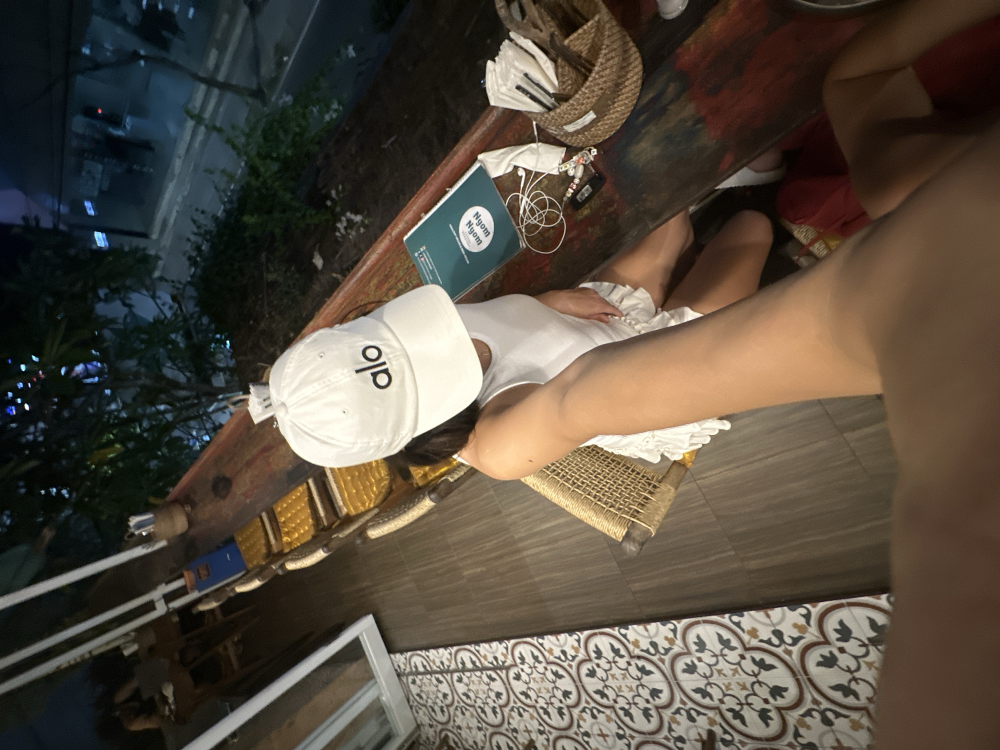 A high-angle shot depicts a person wearing a white "ALO" branded cap and a white top, seated at a weathered, wooden table in what appears to be an outdoor restaurant or patio setting. The location is suggested to be in North Kuta, Indonesia. On the table is a green notebook with the words "NYOM NYOM" printed on it, alongside a pair of white earphones, a small black electronic device, a woven basket with white items inside, and various other items. The table sits atop a patterned tile floor, and in the background, outdoor seating, trees, and a glimpse of a night cityscape are visible. A blurry arm of another individual occupies the right side of the frame, partially obscuring the view.