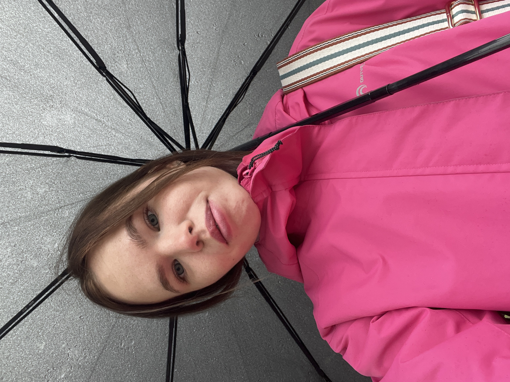 A young woman with light brown hair and blue-grey eyes is captured in a selfie from a low angle, looking directly into the camera with a neutral to slightly playful expression. She is wearing a vibrant fuchsia pink jacket, possibly a rain jacket, which covers most of her torso. A multi-striped strap, featuring brown, white, and teal bands, is visible over her right shoulder, likely belonging to a bag. On the jacket, near this strap, the partial text "OUTVE" and a circular logo are discernible. The woman is positioned beneath an open umbrella, whose black ribs and grey, textured canopy fill the background of the image, suggesting she is outdoors in Chaikovsky, Russia, possibly shielding herself from rain or sun.