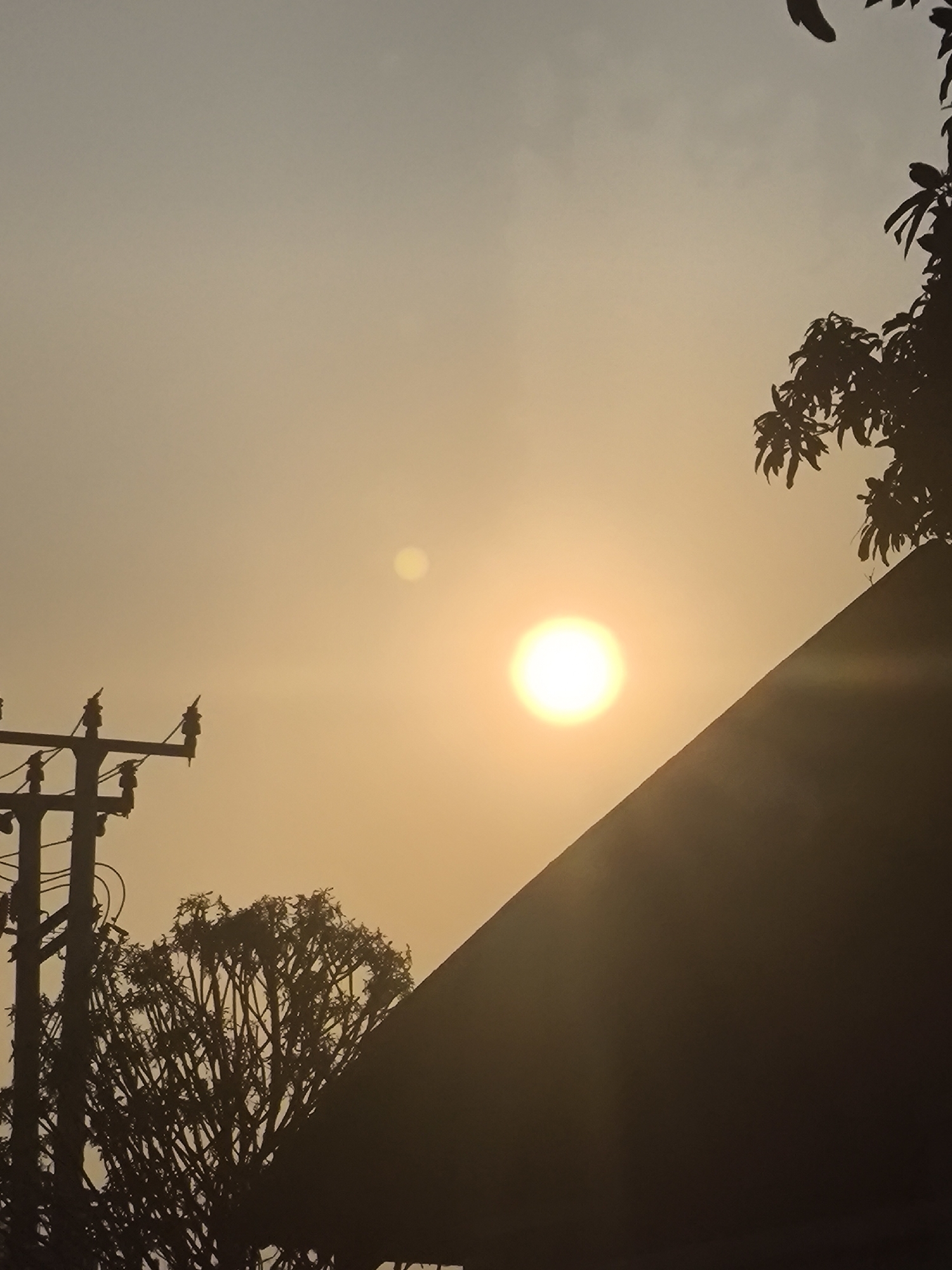 The image captures a sunset in Cilegon, Indonesia, with a hazy, orange sky dominating the frame. In the foreground, a silhouetted power pole with insulators is visible on the left, and to its right, the dense branches of a silhouetted tree. A sharp, dark triangular shape, possibly a building or roofline, cuts across the lower right portion of the image. The bright sun is positioned slightly off-center to the right, appearing as a luminous orb with a faint halo. Another smaller, dimmer orb is visible to the left of the sun. Silhouetted foliage, likely from a tree, is present in the upper right corner. The overall atmosphere is serene and somewhat muted due to the atmospheric conditions.