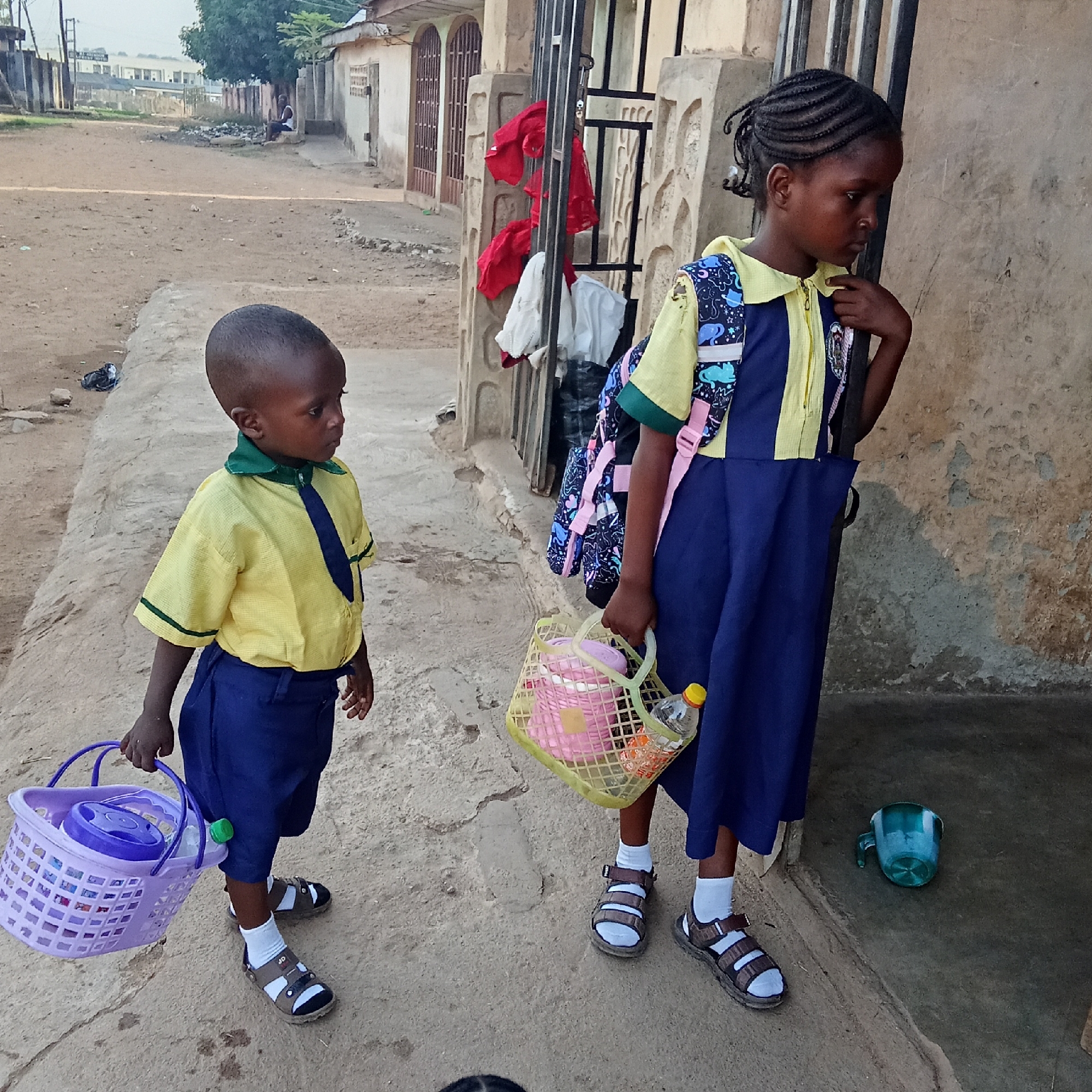Two young children, likely siblings, dressed in school uniforms, stand on an unpaved, dusty path next to a building. The scene suggests a residential area in Akwanga, Nigeria.
On the left, a small boy wears a yellow short-sleeved shirt with a green collar and green trim on the sleeves, blue shorts, a dark blue tie, white socks, and brown and white sandals. He carries a purple plastic basket containing a lidded container.
On the right, a slightly older girl wears a yellow and blue vertically striped dress, similar in color to the boy's uniform. She has a colorful backpack with patterns of blue elephants and other shapes on her back, with pink straps. Her hair is styled in braids. She also wears white socks and brown and white sandals. She carries a light yellow plastic basket holding a pink thermal container and a small plastic bottle with a yellow cap. Her gaze is directed downwards and to her right, giving her a thoughtful or subdued expression. The boy also looks to his right.
In the background, there are concrete buildings with dark metal gates. Red and white clothes are hanging on one of the gates, possibly drying. Further back, a person is seated on a low wall or ledge, and in the distance, a larger building is visible. The ground is rough and appears to be dirt or sand. Near the girl's feet, a green plastic cup lies on the ground. The sandals worn by both children have "JD" visible on the straps. The lighting suggests it might be morning or late afternoon.