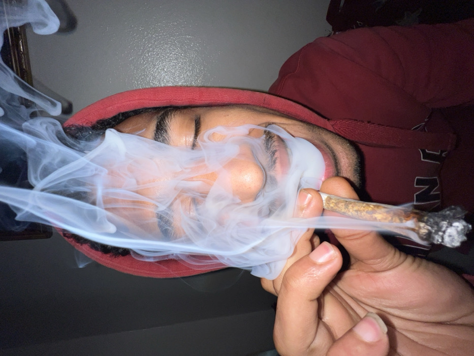 The image portrays a male individual in an indoor setting located in Tanger, Morocco. The subject is wearing a red hooded sweatshirt and is depicted with closed eyes and a subtle smile. The individual's right hand holds a lit, hand-rolled cigarette, from which a substantial plume of white smoke emanates, partially obscuring the lower half of the subject's face. The end of the cigarette exhibits an accumulation of gray ash. The background consists of a plain, light-colored wall, with a darker area to the left and a partially visible dark-framed object positioned in the upper right section. The ambient lighting appears subdued.