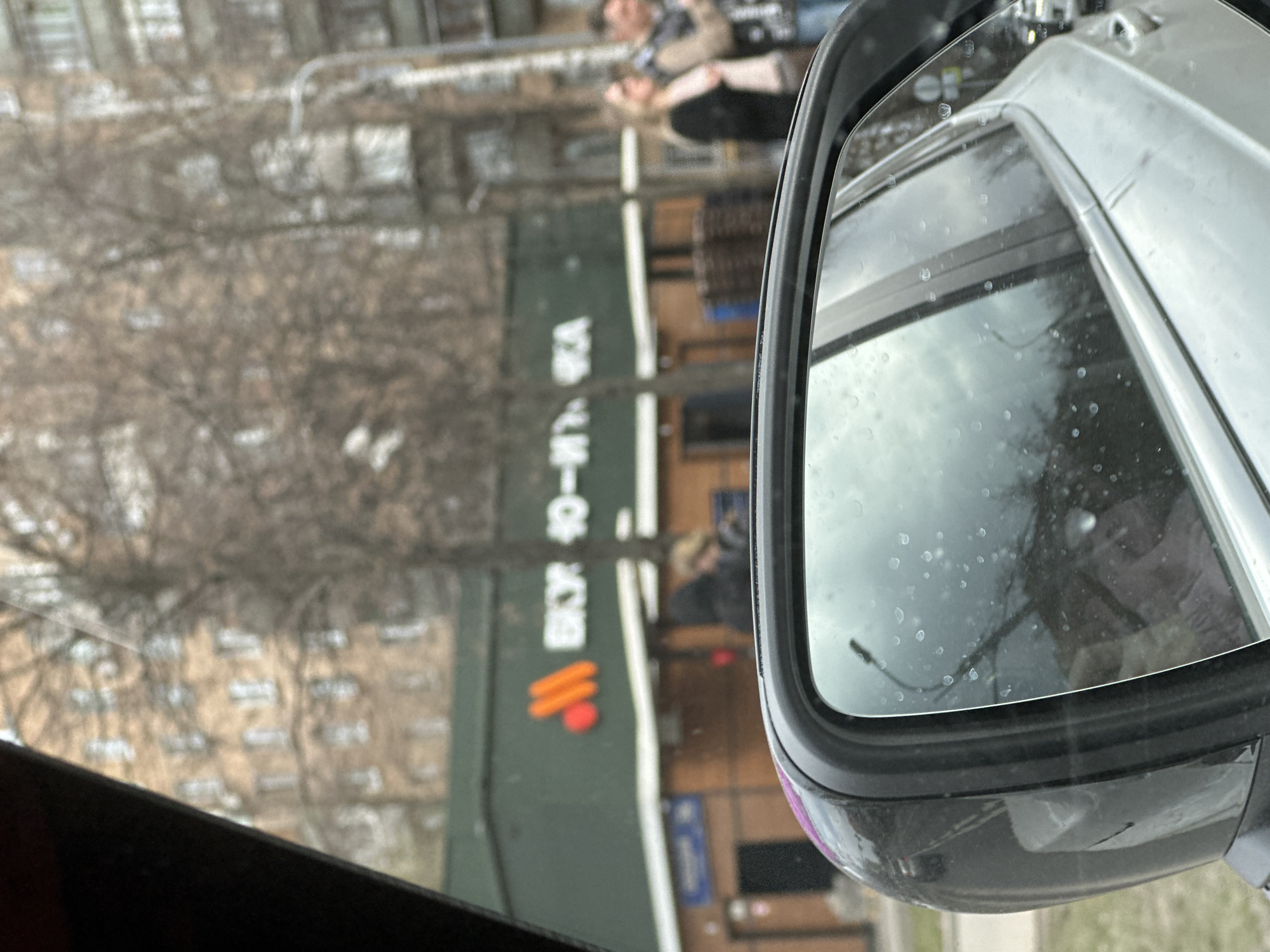The image shows a car's side mirror reflecting a scene in Moscow, Russia. The reflection includes the storefront of "ВКУС - ТОЧКА", a replacement for McDonald's, with its new logo. Bare trees and a building with multiple windows are visible in the background. A blurred individual is walking on the sidewalk. Water droplets are present on the surface of the side mirror.