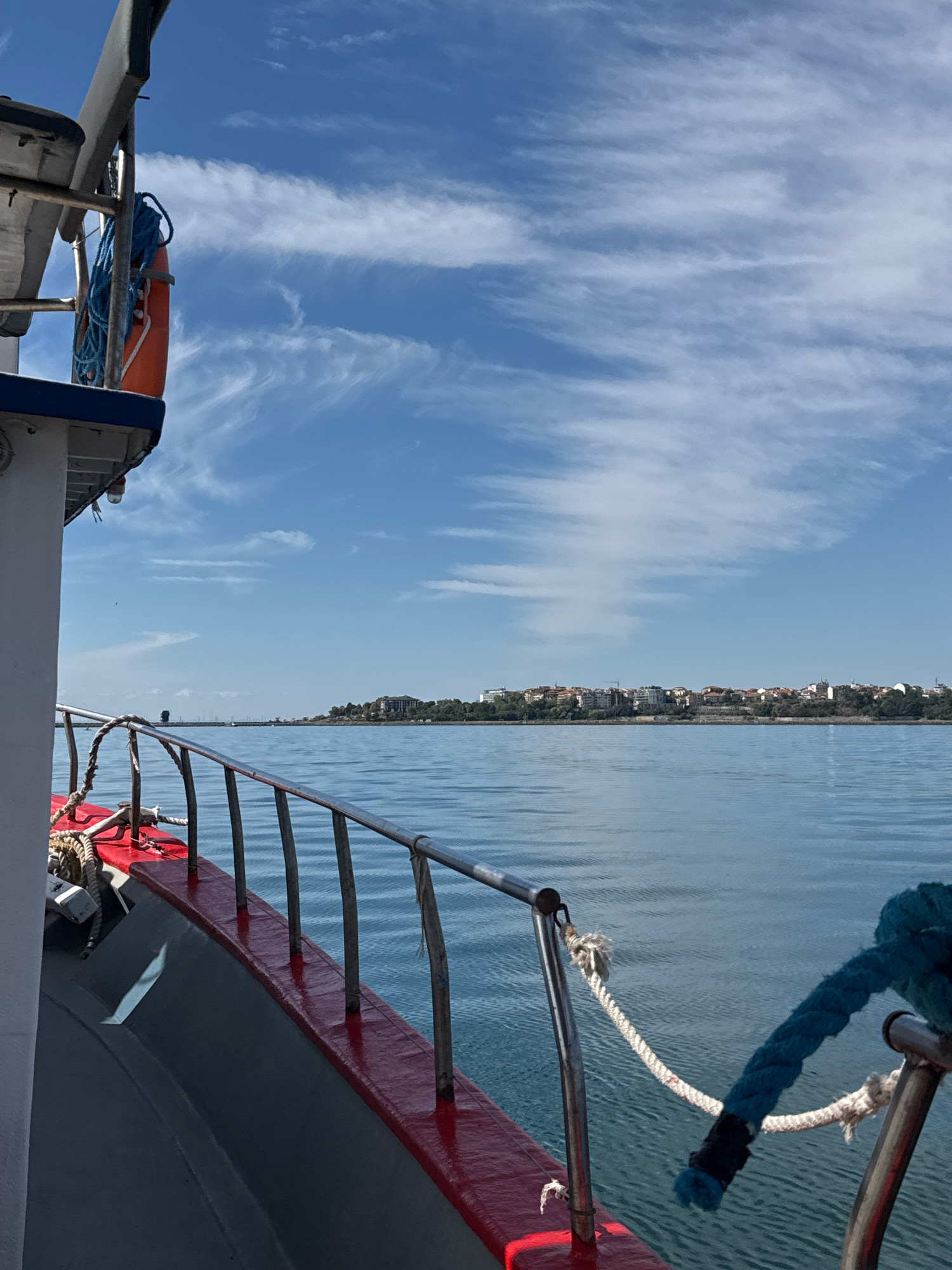 The image shows a view from a boat sailing on the Black Sea towards the ancient town of Nesebar, Bulgaria. The boat's railing, painted red and grey, is visible in the foreground, with a blue rope tied to it. The calm, blue water stretches out to the horizon. In the distance, the historic town of Nesebar is perched on a peninsula, its red-tiled roofs and white buildings clearly visible against the backdrop of lush greenery and a bright blue sky dotted with wispy clouds. The scene captures a serene moment of travel, offering a picturesque glimpse of Nesebar's coastal landscape.