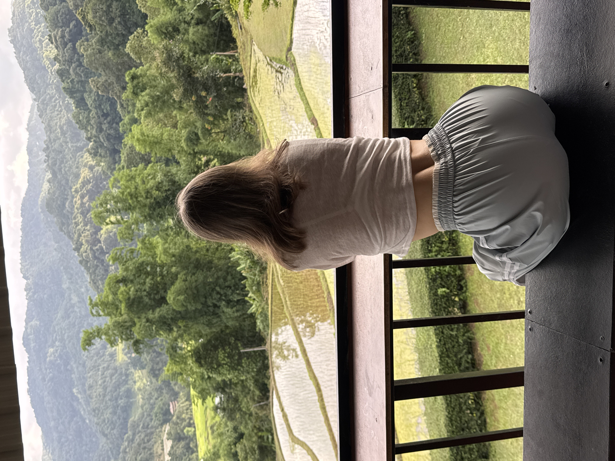 A young woman with brown hair is seated on a dark surface, facing away from the camera and looking out at a scenic view. She is wearing a light-colored t-shirt and light blue patterned pants. The background features lush green rice terraces, dense foliage, and rolling hills under a cloudy sky, indicative of a rural mountainous region. The scene appears to be from a balcony or elevated vantage point, with a wooden railing in the foreground. The location is identified as Ban Ang Ka Noi, Thailand.