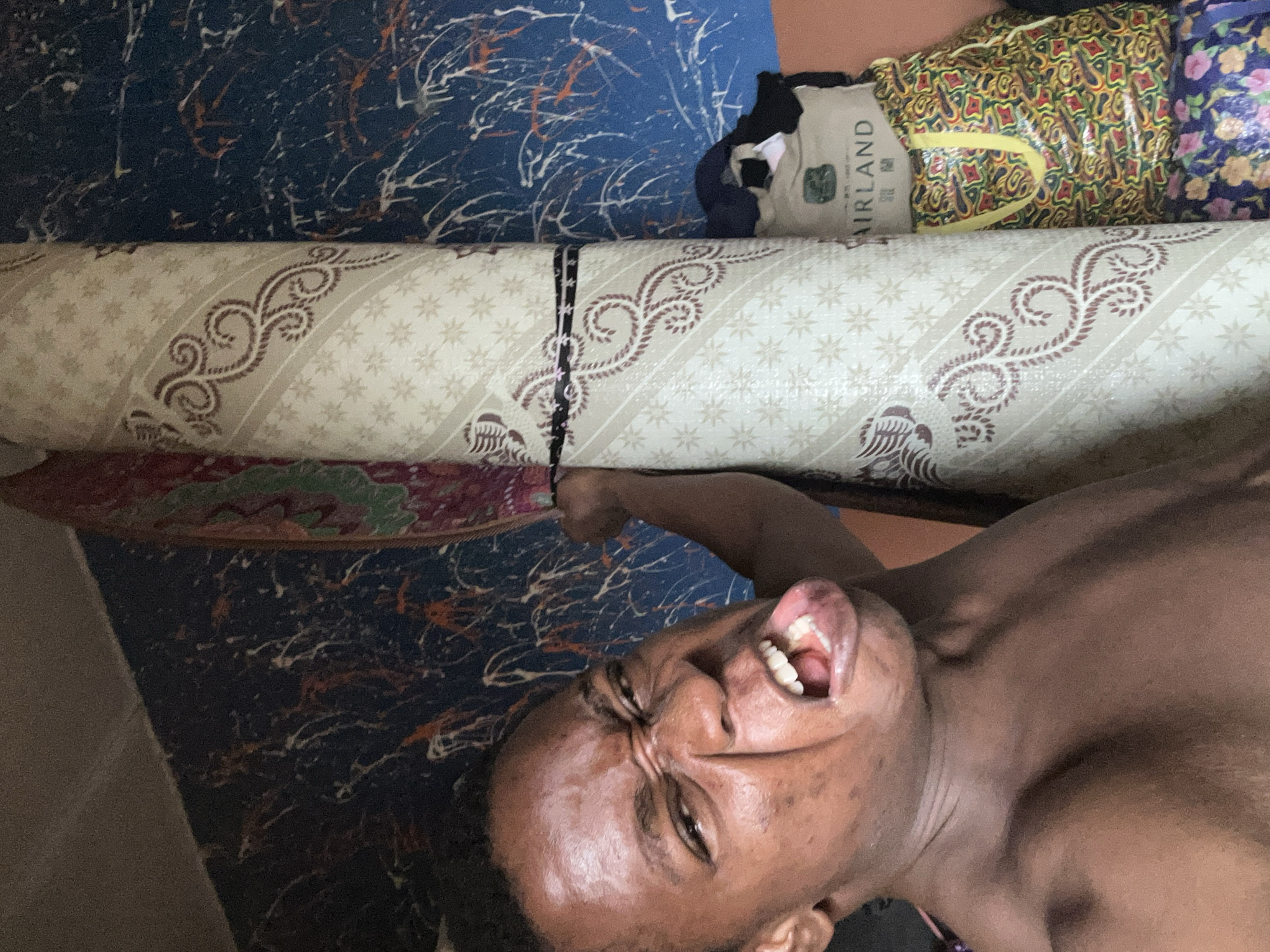 The image shows a person with dark skin, eyes closed, and mouth open with their tongue partially protruding. The person is lying on what appears to be a rolled-up rug or mat with a patterned design. Behind the roll is a blue surface with white and orange markings, possibly another rug or a decorated floor. A shopping bag and other bundled items are visible in the upper right corner. The location is described as Unknown, Unknown.