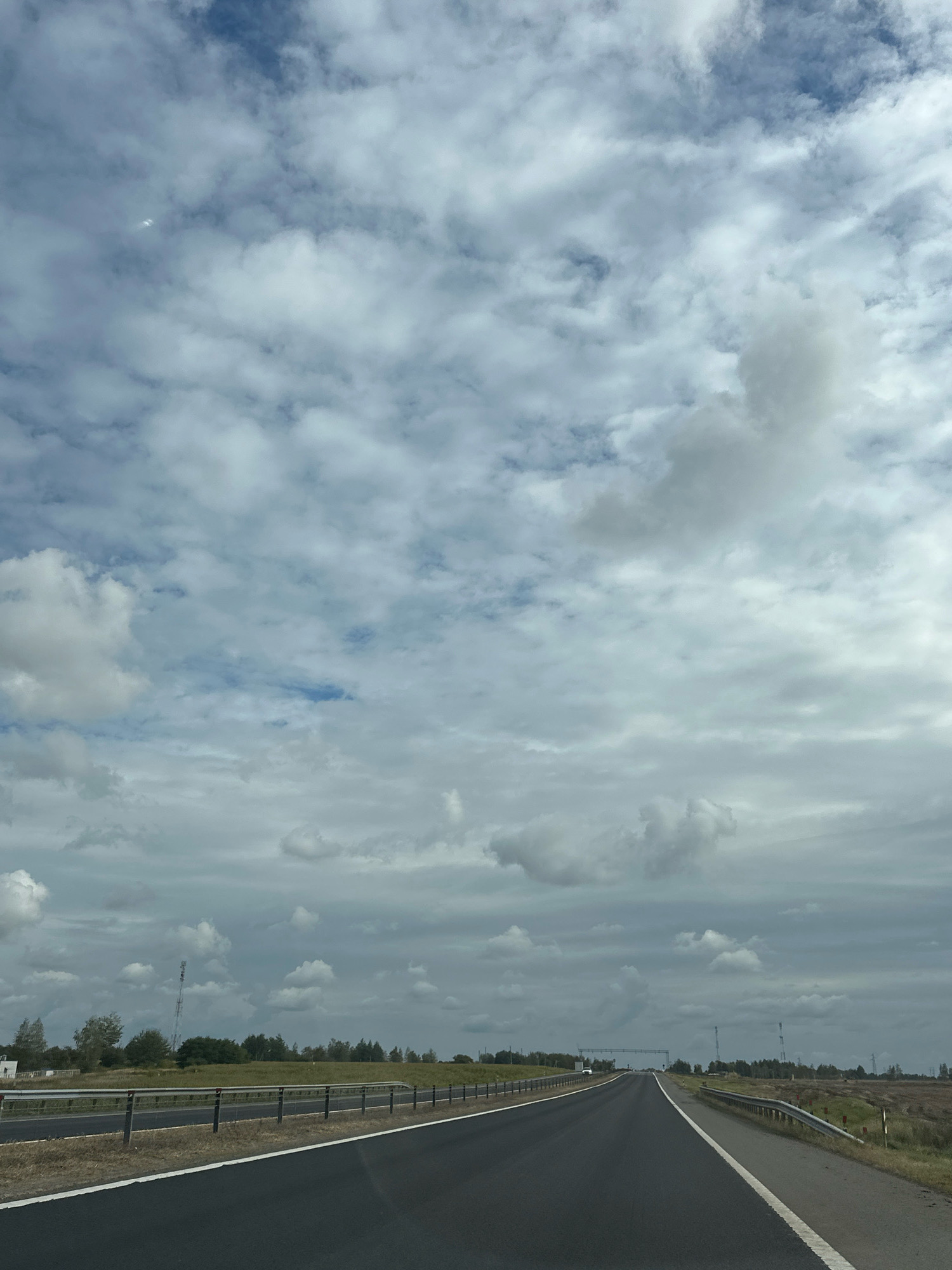 A straight, paved road with white lane markings extends into the distance under a cloudy sky. The road is bordered by metal guardrails and a grassy shoulder. In the background, there is a flat, open landscape with trees and a few structures, including what appear to be communication towers. The sky is filled with a mix of white and gray clouds, with patches of blue sky visible. The overall impression is of a rural or semi-rural highway. The image appears to be taken from a vehicle moving along the road.