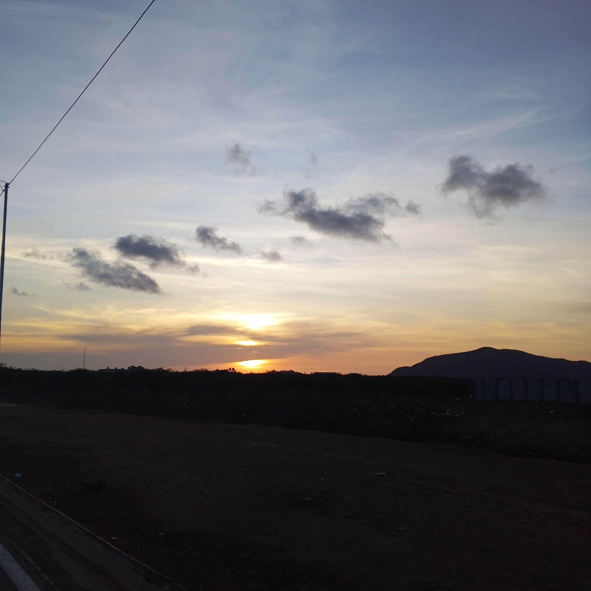 The image depicts a sunset in Unknown, Unknown. The sky is a blend of blue, white, and orange hues, with scattered grey clouds. The sun is setting behind a dark, silhouetted landscape with a hill in the background and a fence in the midground. The foreground is a dark, dirt surface. A street lamp is visible on the left, and a power line stretches across the upper part of the image.