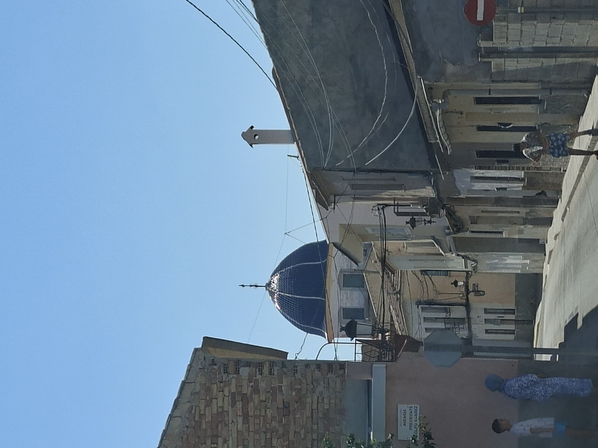 The image depicts a street scene in Granja De Rocamora, Spain, dominated by a bright blue domed building visible in the background. The street is lined with buildings of varying architectural styles and states of repair. On the left side, a brick building is partially visible. In the foreground, two individuals are standing on the sidewalk next to a street sign. One is a child wearing a white t-shirt and blue shorts, and the other is a person wearing a blue head covering and a patterned blue and white outfit. Further down the street, another person is standing, wearing a patterned shirt and blue shorts. Overhead, power lines crisscross the sky. A red and white circular sign indicating a prohibition is visible on the right side of the street. The sky is clear and blue, suggesting a sunny day. The overall impression is of a quiet, sun-drenched street in a Spanish town.