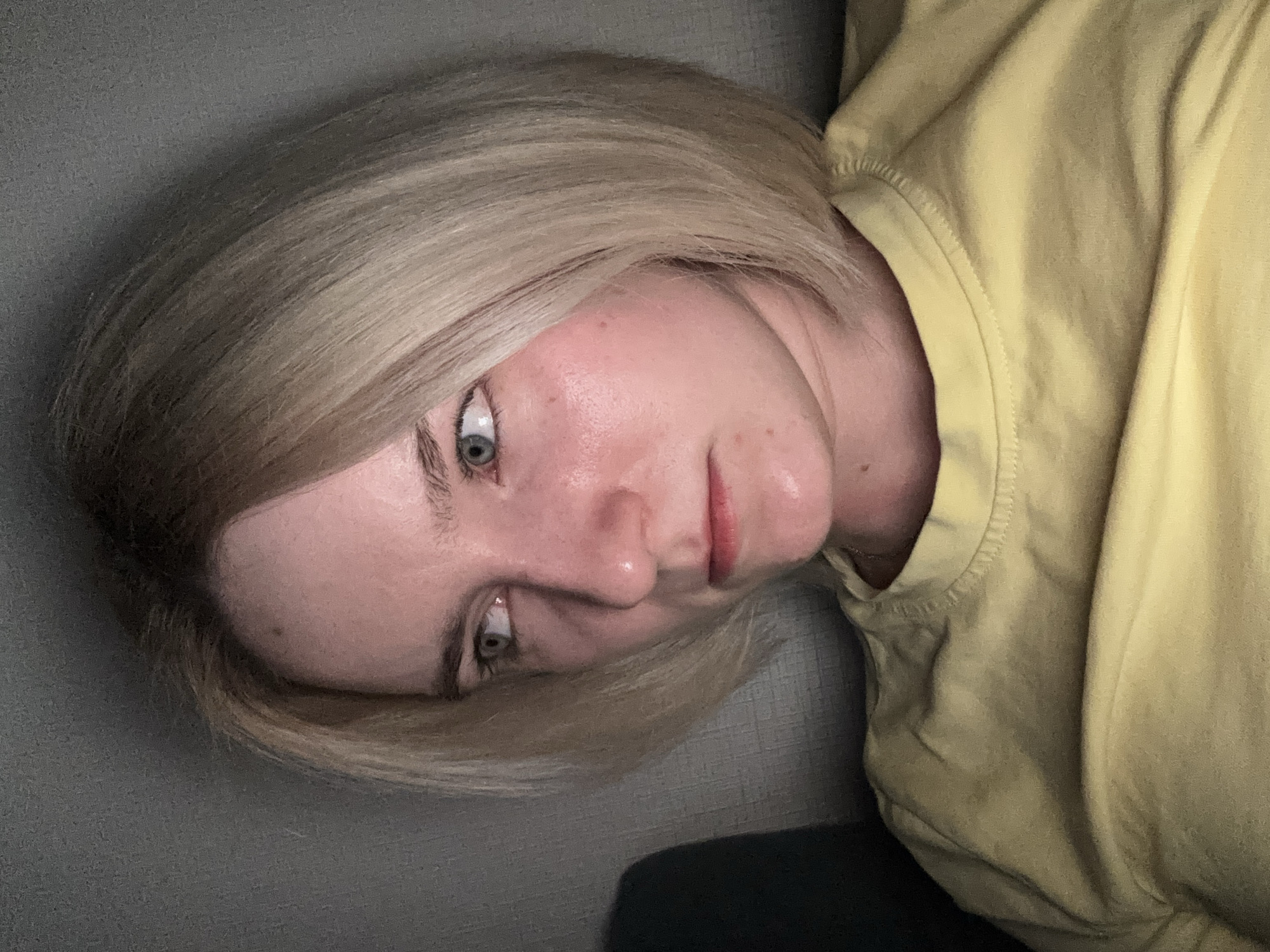 A woman with short, blonde hair is lying on a gray surface, possibly a couch. She is wearing a yellow t-shirt. The image is a close-up, focusing on her face and upper torso. There is no information in the image to determine a location in Vsevolozhsk, Russia.