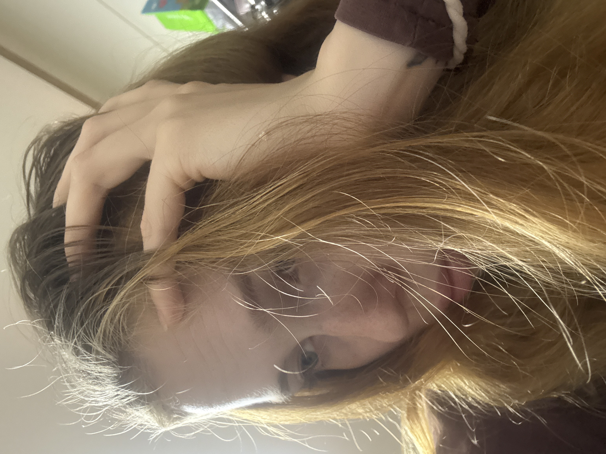 A close-up, low-angle shot shows a person's head and hand, with the image oriented sideways. The person has long, blonde hair with some darker roots visible. Their left hand is in their hair, with fingers spread and partially obscuring their forehead and scalp. The person's face is visible, with their eyes closed and their mouth slightly open, revealing their tongue. Their skin is pale, and there are some wisps of hair falling across their face. They are wearing a dark-colored garment, possibly a sweatshirt or long-sleeved shirt, with a white trim visible on the neckline on the left side of the frame. The background is blurred but appears to be an indoor setting with some lighter-colored objects. The lighting suggests it might be daytime, possibly indoors with artificial light. The overall mood appears relaxed or introspective. No visible text is present in the image.