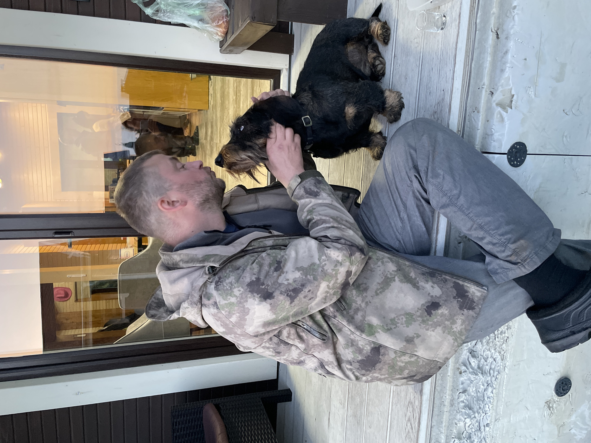 A man sits on a porch or patio, facing left, and is petting a black and brown terrier-type dog. The man is wearing gray pants, black socks, and black boots. He has on a camouflage jacket over a gray top. He is fair-skinned with short blond hair and a beard. He is looking at the dog and has his hand on its head. The dog is standing on the porch or patio and looking at the man. In the background, there is a large glass window reflecting interior objects, including furniture and people. Above the man and the dog is a dark wooden shelf with a plastic bag on it. The porch or patio appears to be made of light-colored wood and has some peeling paint in the lower right corner. The location is likely outdoors, as the lighting suggests natural daylight. There is no visible indication of Solov'evka, Russia.