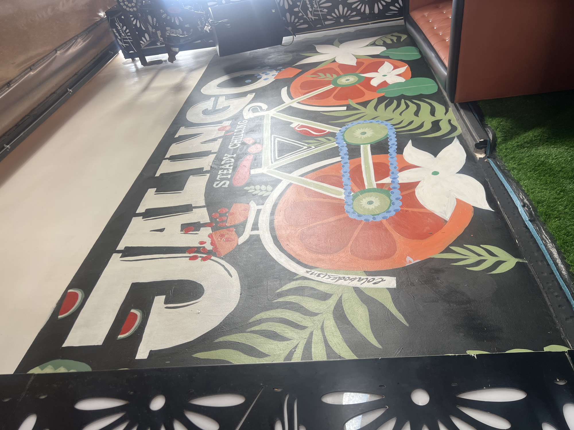 The image shows a floor mural in a dimly lit indoor setting. The mural is painted on a dark surface and features large, stylized lettering that reads "GO FIND". Interspersed with the lettering are tropical-themed illustrations, including sliced oranges, white flowers, green leaves, and a blue and green beaded necklace. The style is vibrant and playful, suggesting a relaxed or entertainment-oriented location.
In the background, a portion of a booth with brown upholstery and tufted details is visible, alongside a strip of artificial green turf. To the left, a metallic object with a decorative pattern, possibly a fan or a light fixture, is partially visible. The overall impression is that of an indoor space designed for leisure or dining, with a tropical or beachy theme. The lighting appears artificial, casting shadows and creating a somewhat moody atmosphere. The presence of the booth seating and the artificial turf suggests this could be a bar, restaurant, or lounge. No people are visible in the image.
There is a small, handwritten label within the mural that reads "Colaborates".