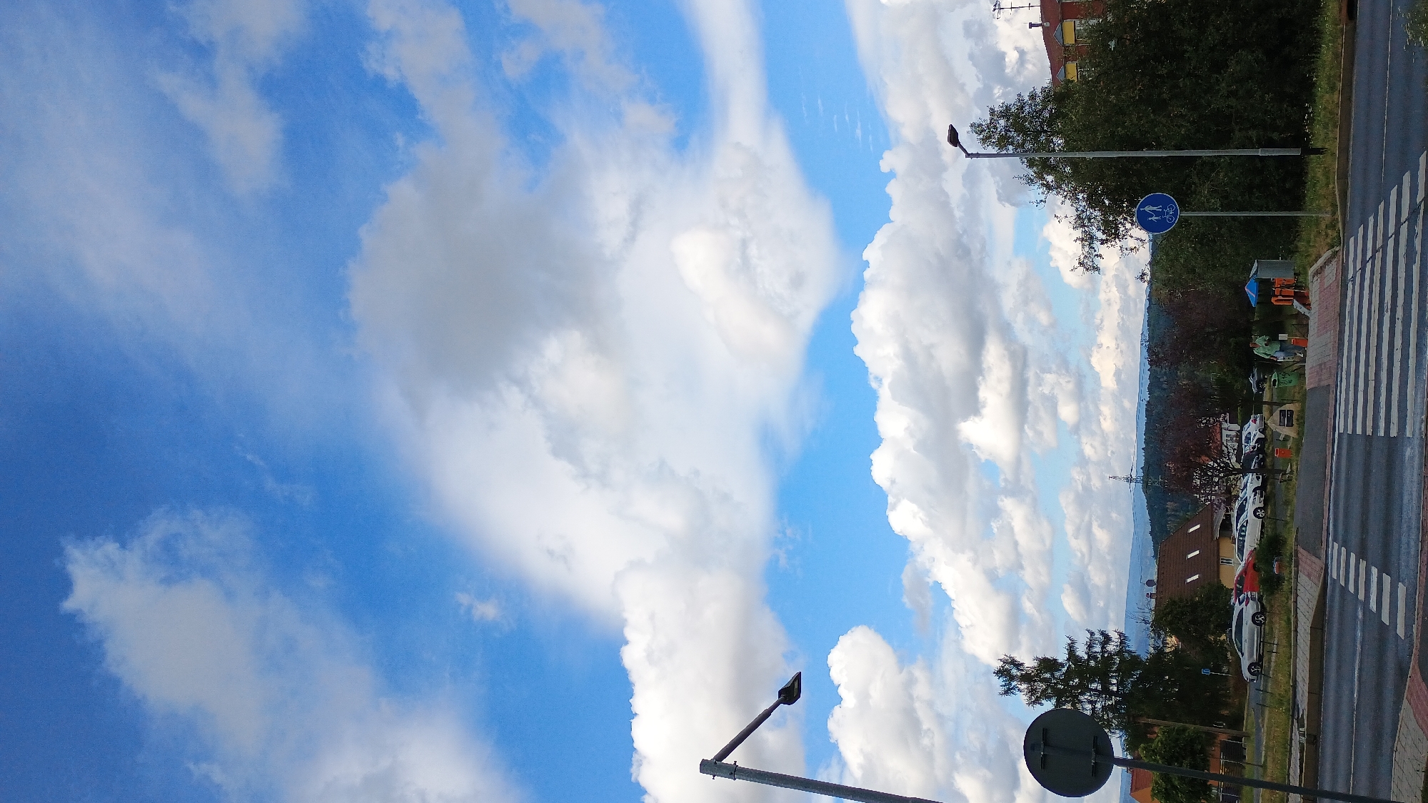 A street scene in Sokolov, Czechia, captured on a cloudy day. A pedestrian crossing is visible in the foreground. Beyond the crossing, there are parked cars, trees, and buildings. A traffic light and a bicycle path sign are also present. The sky is filled with white clouds against a blue backdrop.