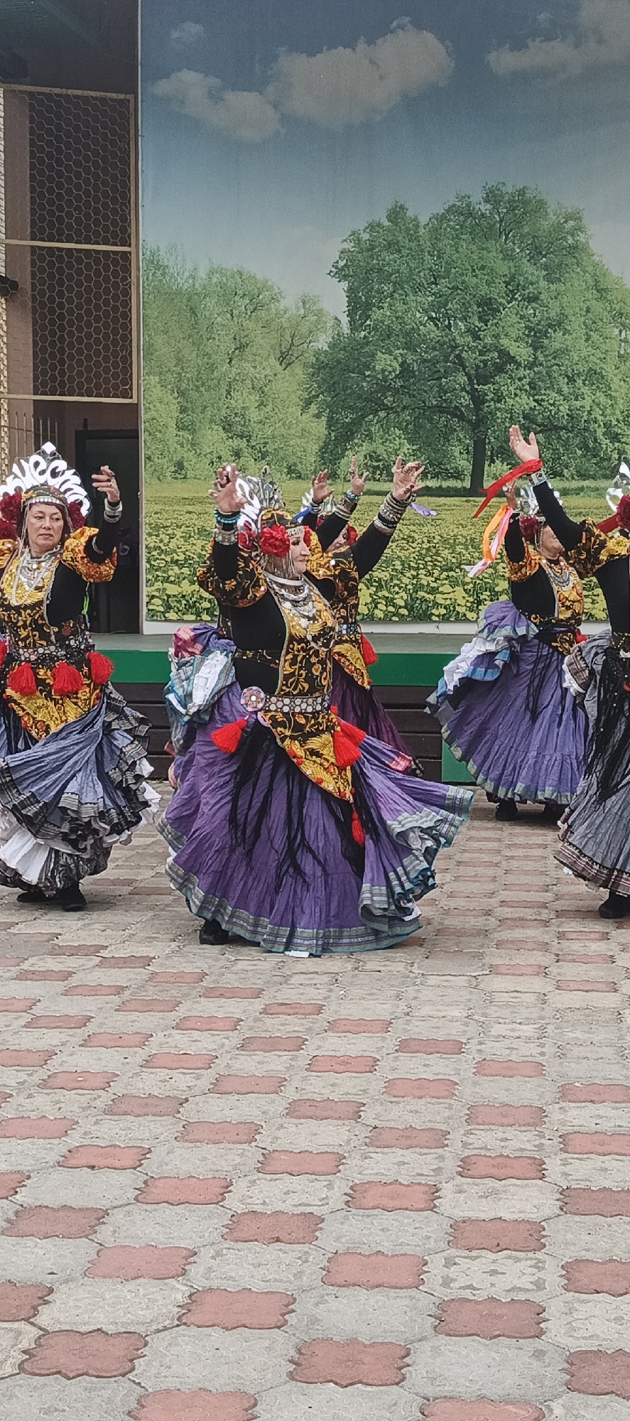 This image captures a vibrant cultural performance featuring a group of women dressed in elaborate, colorful traditional attire. They are engaged in a lively dance, their full skirts and ornate headdresses adding to the visual spectacle. The costumes are a mix of rich fabrics, intricate patterns, and bold embellishments, including red tassels and layered jewelry.
The setting appears to be outdoors, with a brick-patterned pavement in the foreground. In the background, a large mural or backdrop depicts a picturesque landscape with green trees and a blue sky with clouds, creating an artistic and somewhat artificial setting for the performance. To the left of the mural, a section of a building with a wire mesh pattern is visible.
The women's postures and hand gestures suggest energetic movement, indicative of a traditional dance. Their expressions, though somewhat obscured by the distance and motion, seem focused and engaged in the performance. The time of day appears to be daytime, given the bright lighting. The weather seems fair, suitable for an outdoor event.
The overall impression is one of cultural celebration and artistic expression. The rich costumes and dynamic dance movements are the focal points, set against a backdrop that aims to evoke a natural, scenic environment. There is no visible text in the image. The location context provided is Chaikovsky, Russia.