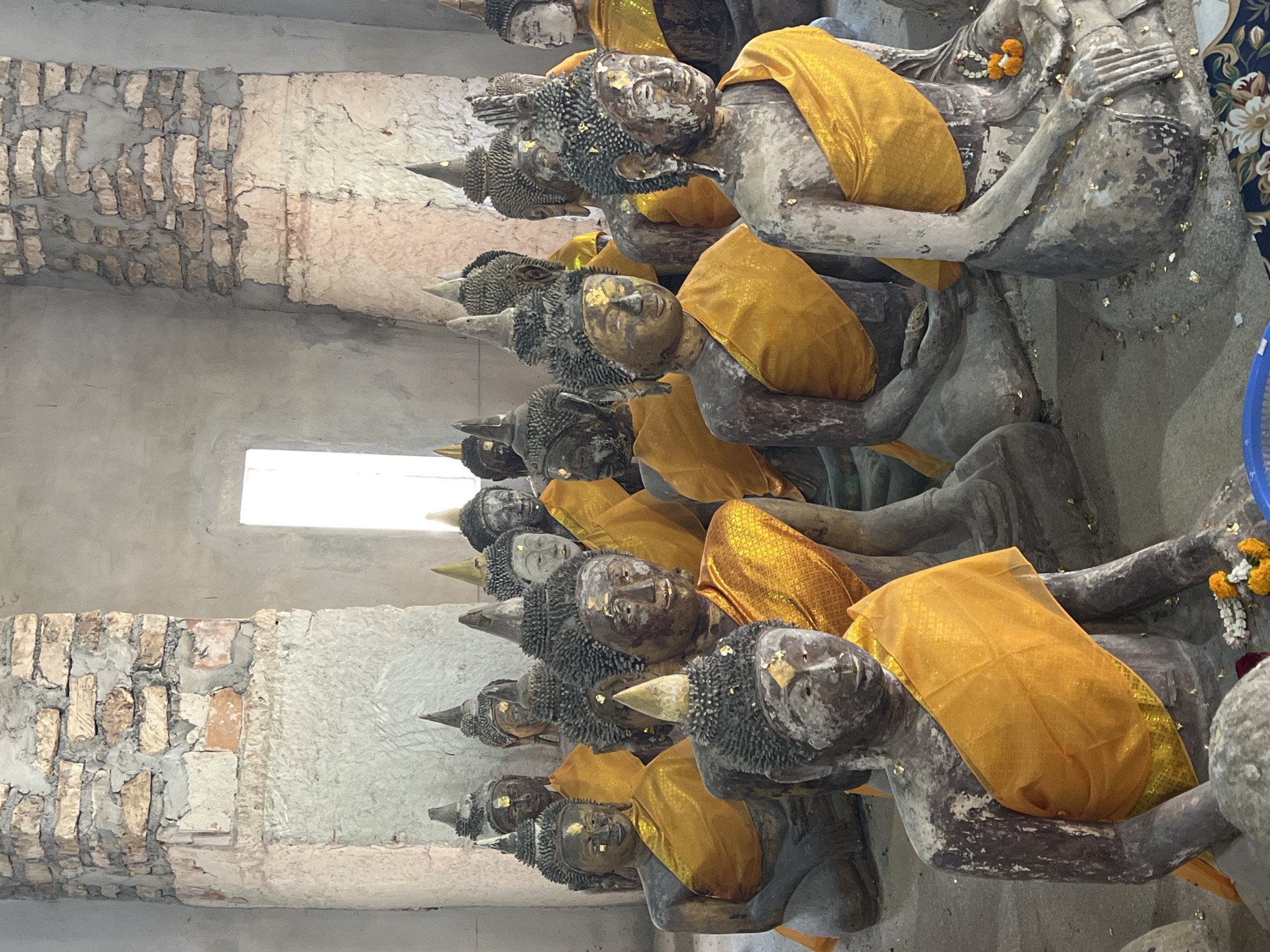 The image depicts a collection of ancient, weathered Buddha statues seated in rows within an old, unfinished building. The statues, made of a dark, rough material, are adorned with saffron robes and have touches of gold leaf on their faces. The building's walls are constructed of rough stone and concrete, with exposed brickwork in some areas. A rectangular opening in the back wall allows light to enter, illuminating the dusty interior. The statues are arranged closely together, some with their heads or shoulders overlapping. The floor is covered in sand and debris, with a few scattered offerings of flowers and leaves near the statues. The scene suggests a place of worship or historical significance, likely a temple or shrine in a rural or historical setting. The overall atmosphere is one of age, reverence, and quiet contemplation.