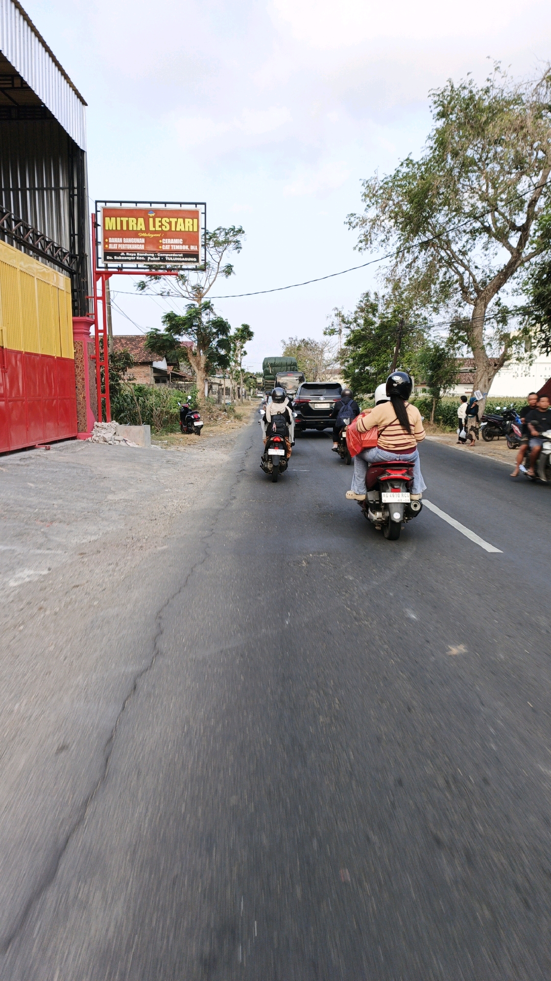 An exterior shot captures a road in Sukoanyar, Indonesia, with a focus on traffic and roadside businesses. A prominent sign for "MITRA LESTARI" advertising building materials, ceramics, and paint stands on the left side of the road, adjacent to a building with red and yellow siding. Several motorcycles and a black SUV are on the road, with some individuals riding on the motorcycles. Trees and houses line the background, suggesting a semi-urban or rural setting. The sky is partly cloudy.